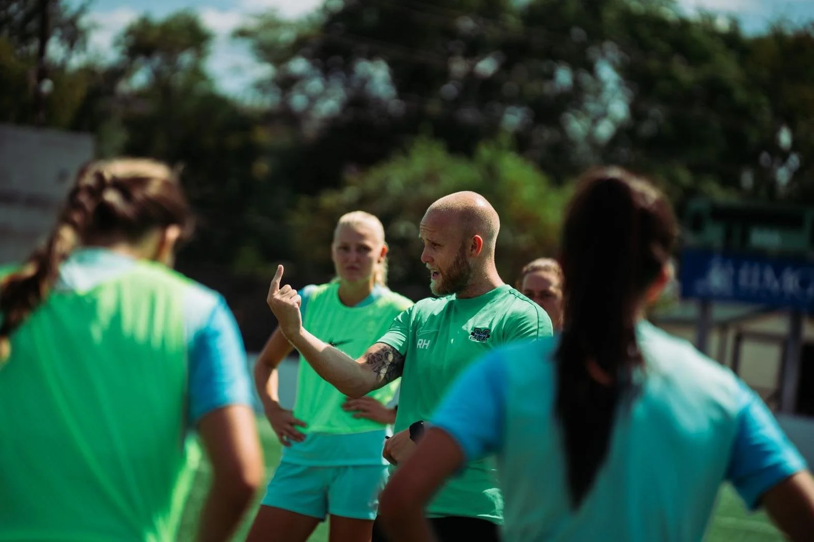 A coach in a green shirt giving instructions to female soccer players on the field, with trees and a sports facility in the background.