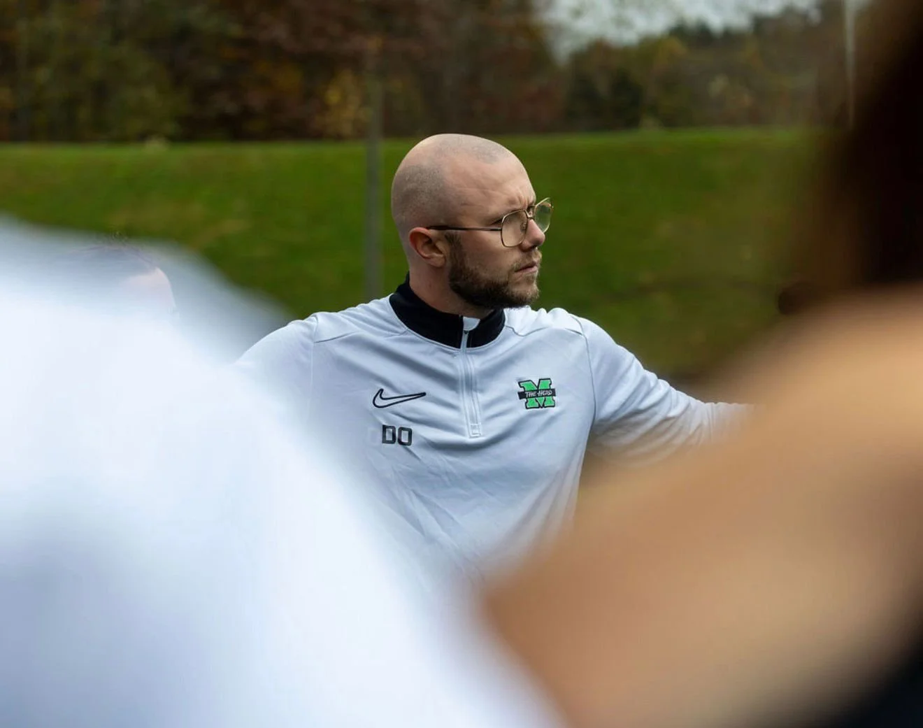 A man with a beard, glasses, and a shaved head, wearing a white sports jacket with green and black logos, standing outdoors with blurred greenery in the background.