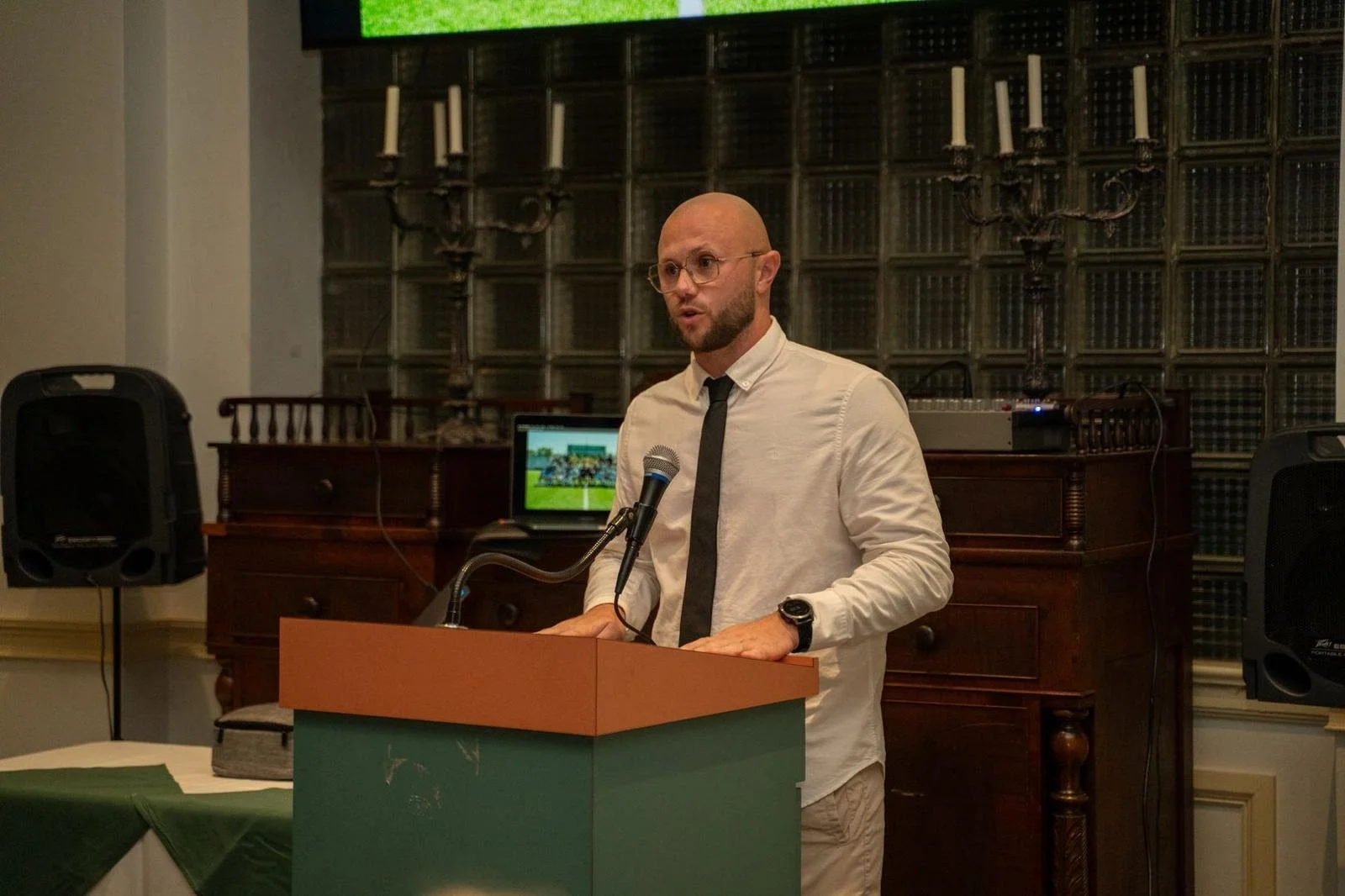 A man in a white shirt and black tie giving a speech at a podium in a room with dark wood furniture and a glass block wall.