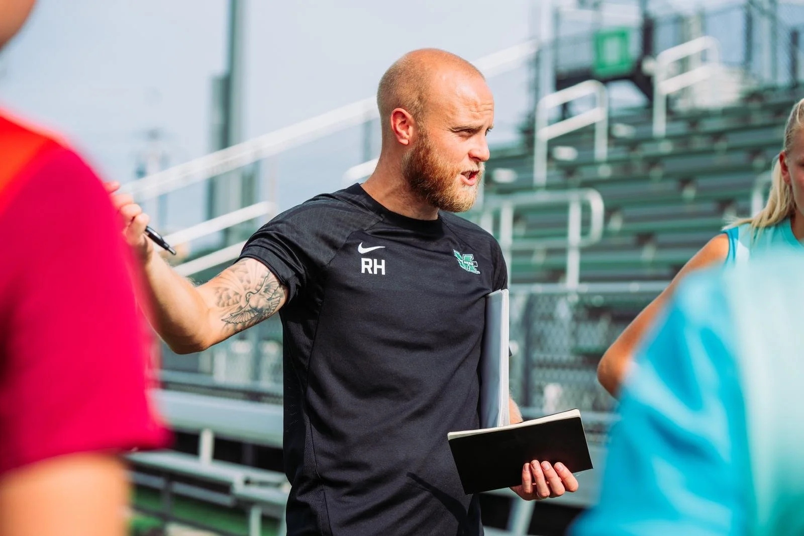 A coach with a beard, tattoos, and wearing a black sports shirt with the letters 'RH' and a logo, stands with a clipboard, speaking to athletes on a sports field with bleachers in the background.