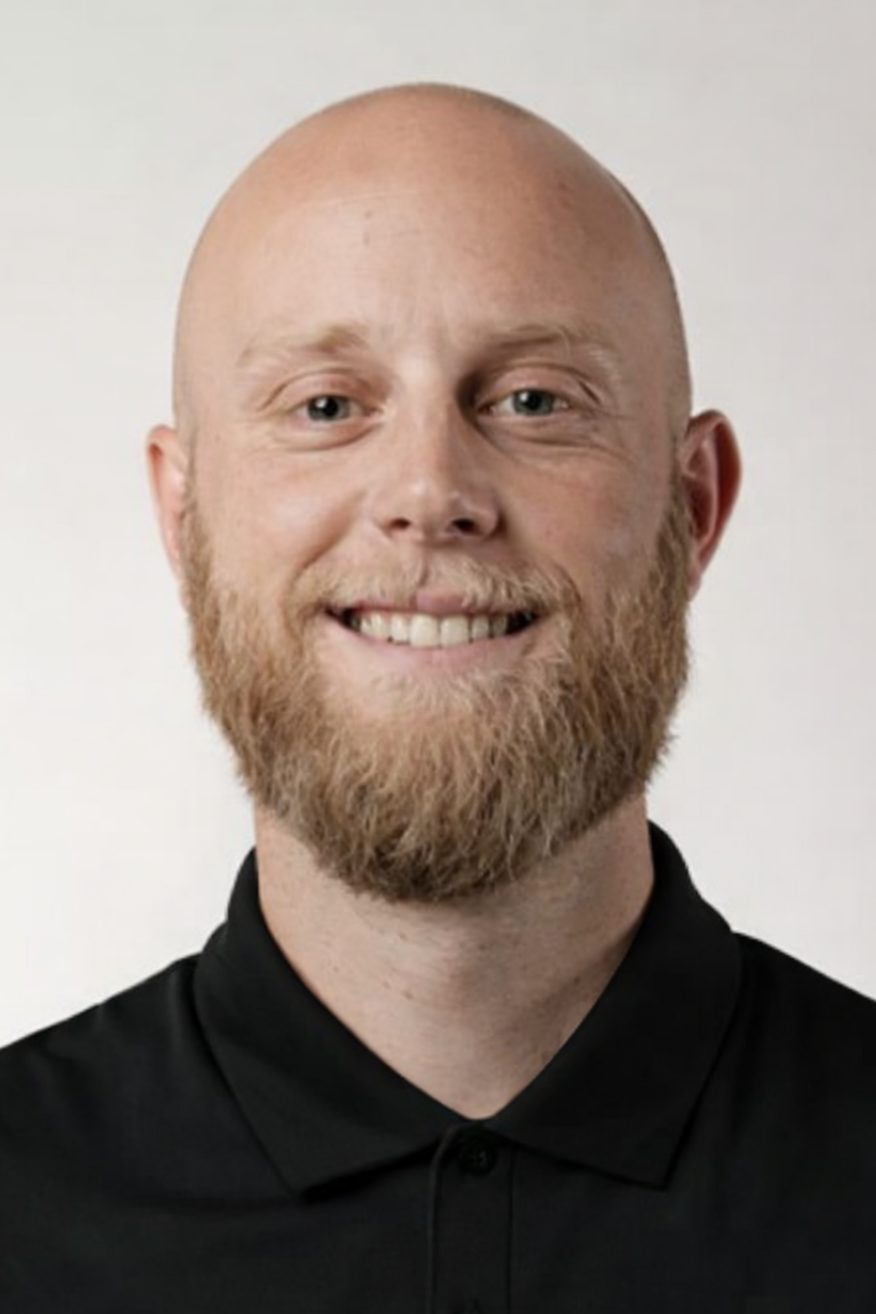 A smiling man with a bald head and beard, wearing a black collared shirt, standing against a plain light background.