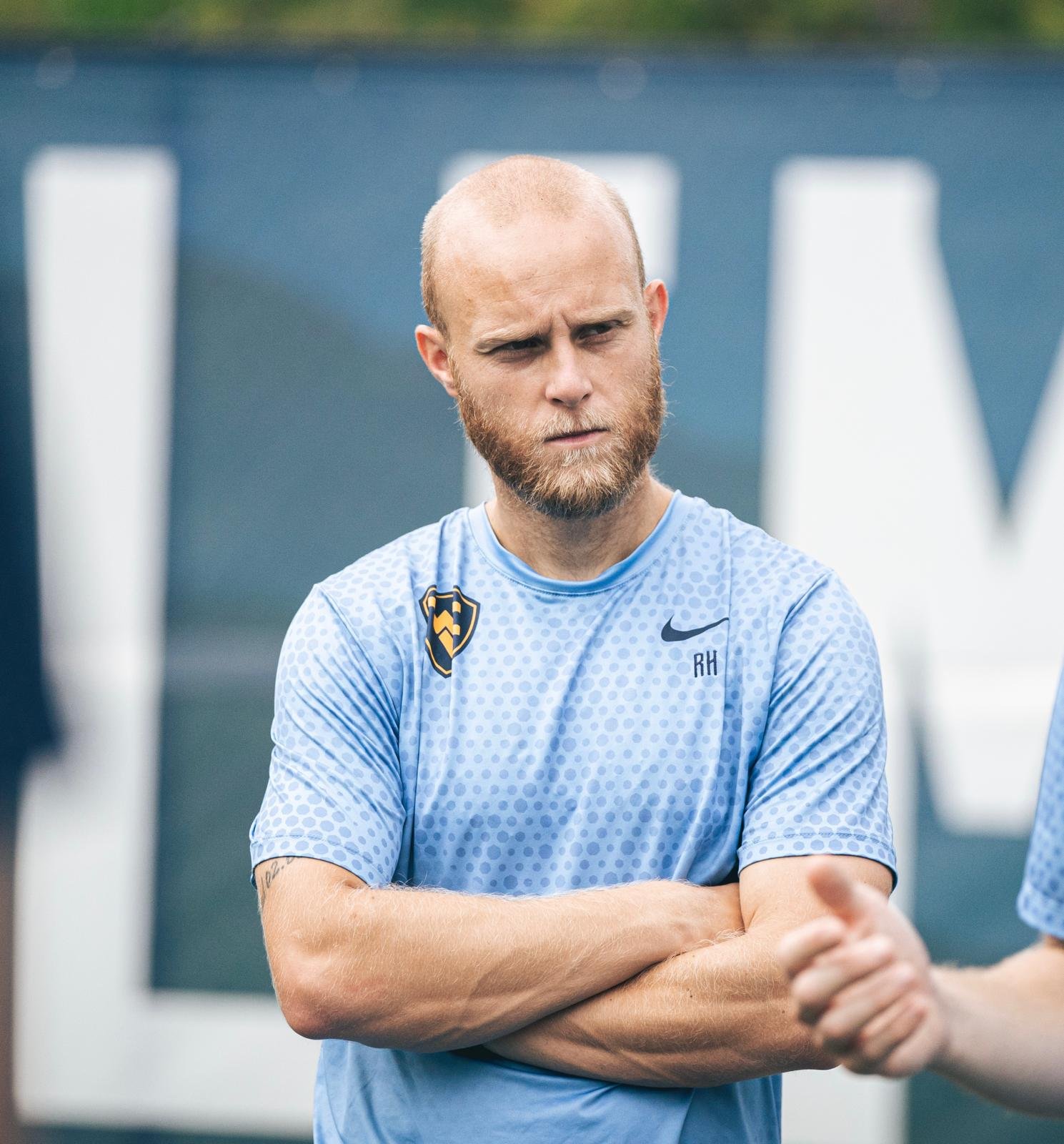 A man with a beard and short hair wearing a light blue sports jersey with a black logo on the chest, standing outdoors with arms crossed.