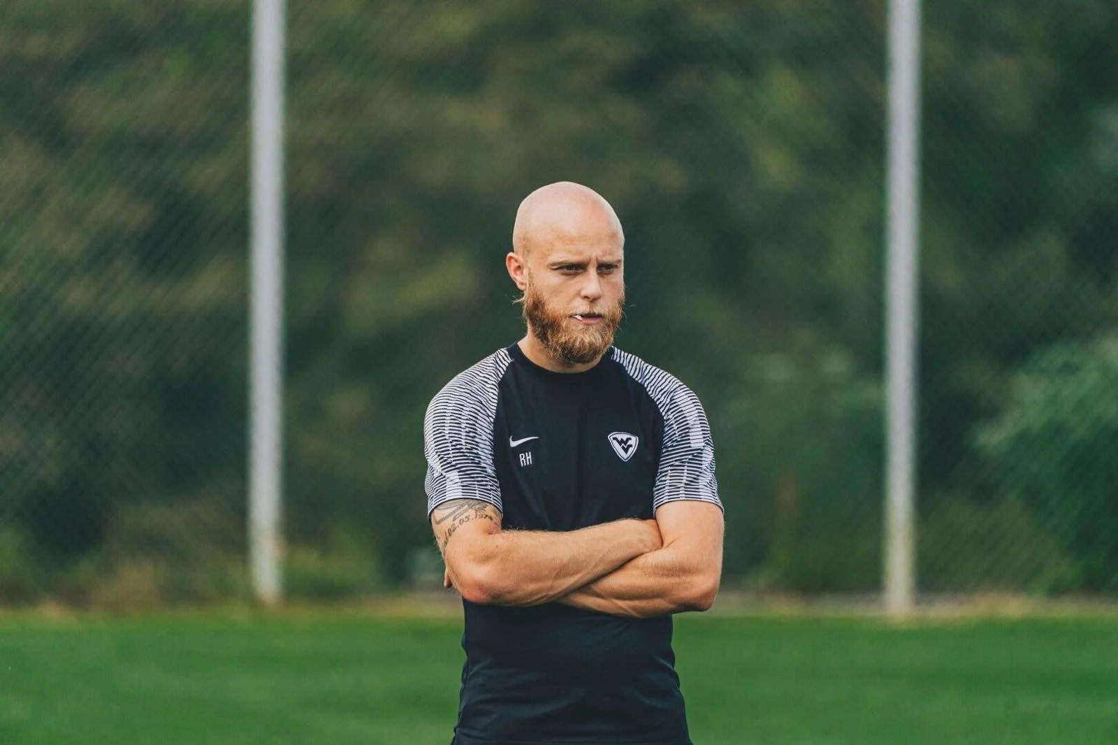 A man with a shaved head and beard stands on a soccer field with arms crossed, wearing a black athletic shirt with a logo and the initials 'RH' on it. The background shows a green fence and trees.