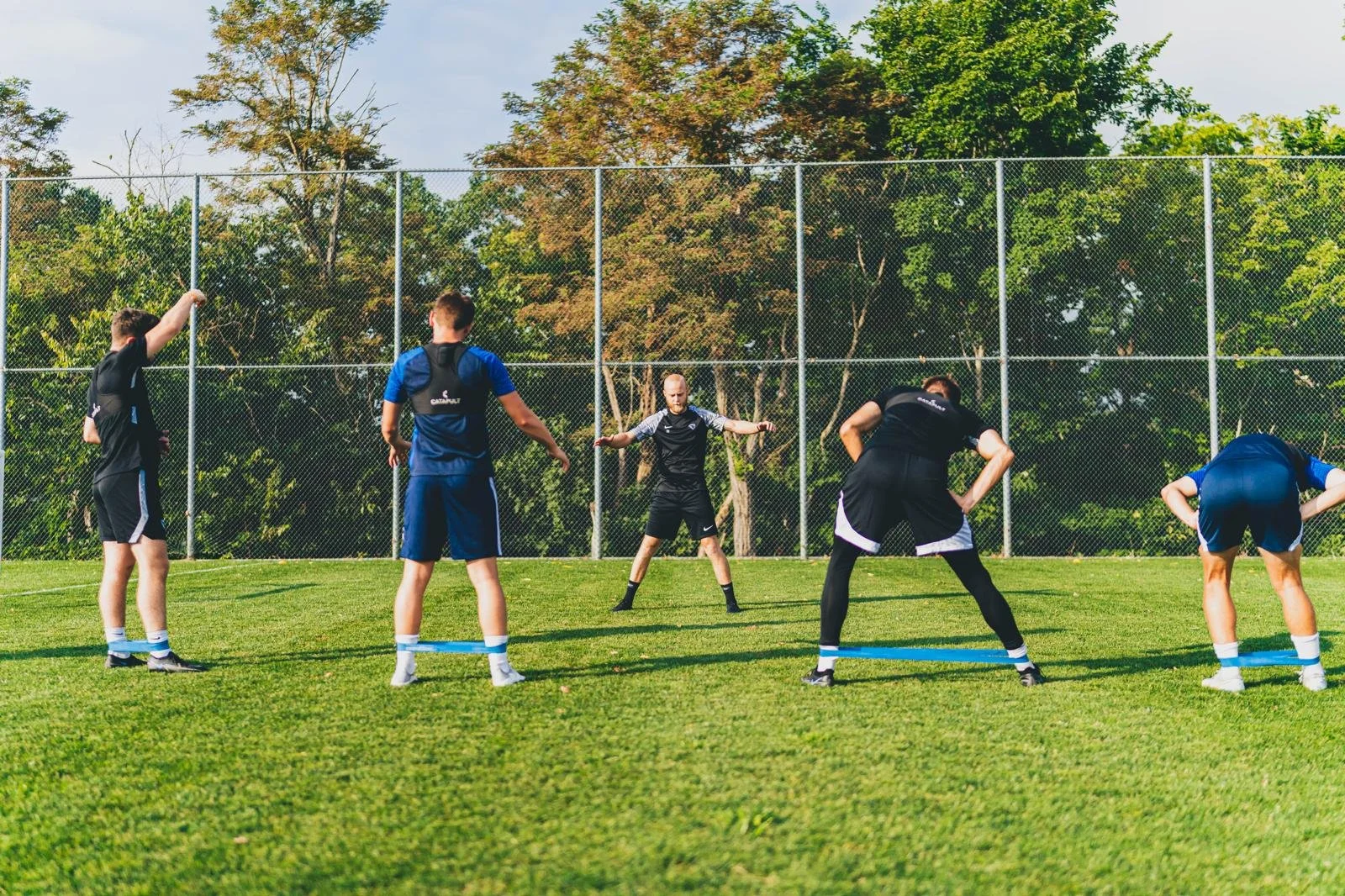 Group of people exercising outdoors on a grassy field with resistance bands, surrounded by trees and a chain-link fence.