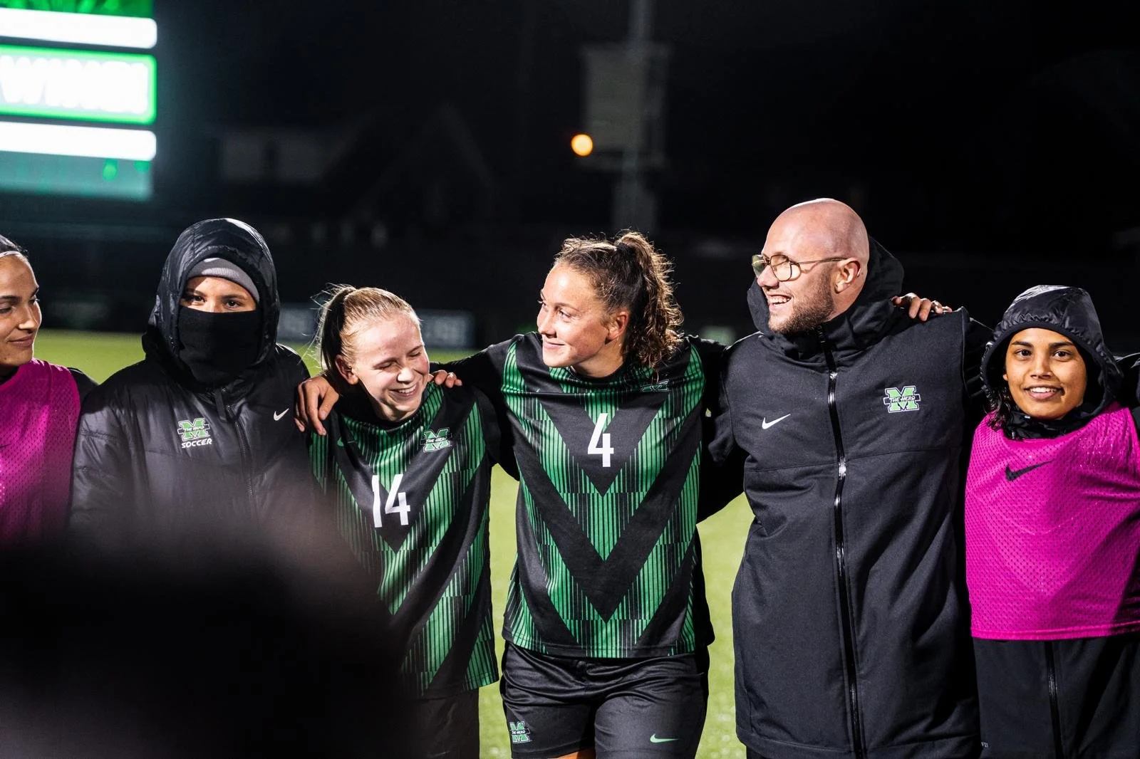 Group of female soccer players and coaches standing on a soccer field at night, smiling, with their arms around each other, wearing team jerseys and jackets.
