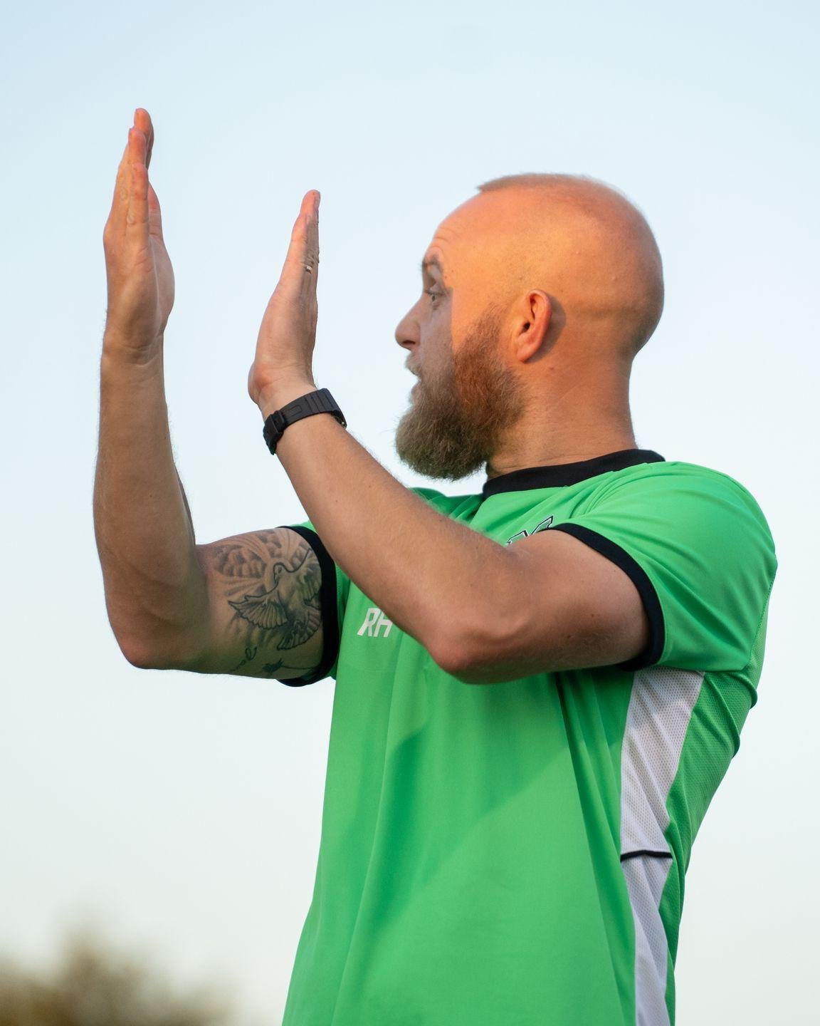 A man with a bald head and beard, wearing a green sports jersey, raising both hands in a stopping or signaling gesture outdoors against a clear sky.