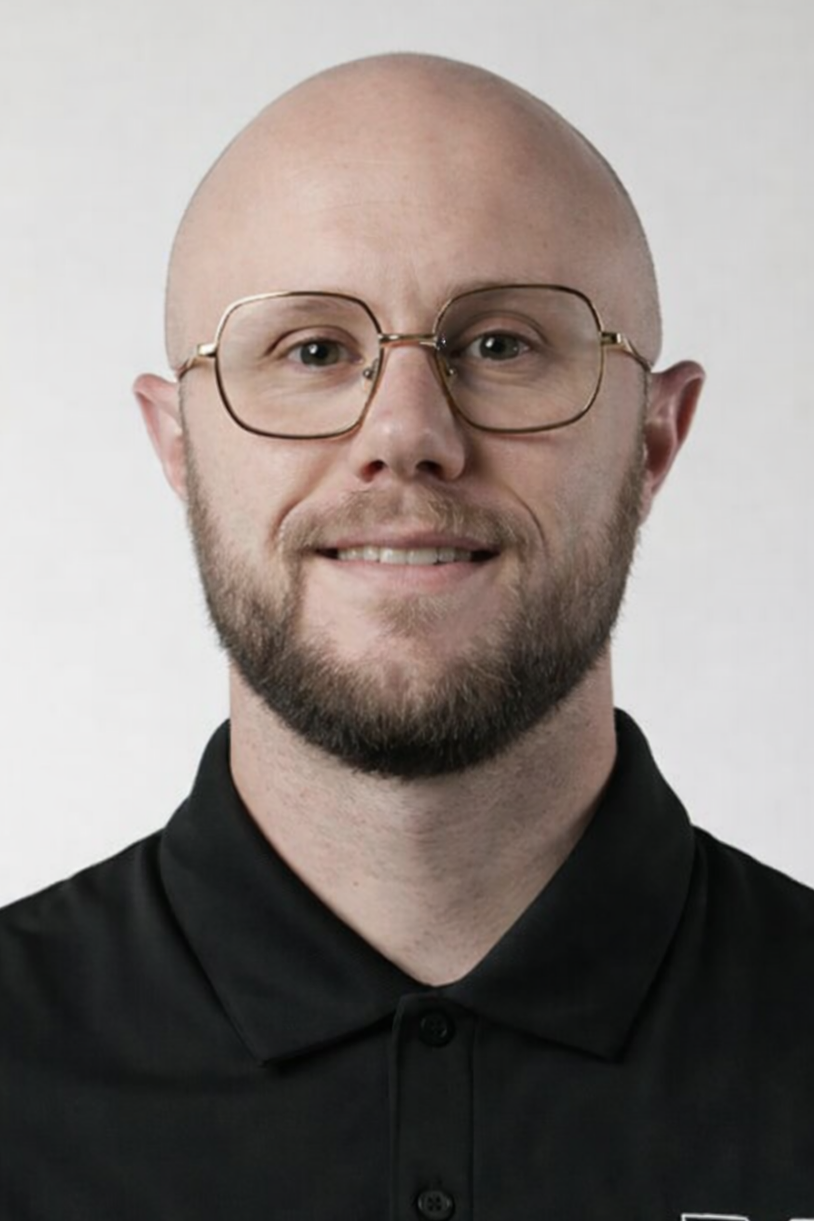 A man with a shaved head, glasses, and a beard, wearing a black collared shirt, smiling at the camera against a plain light background.