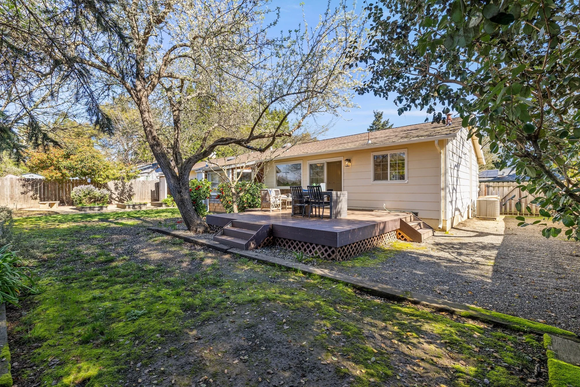Backyard with a wooden deck, outdoor furniture, trees, and a gravel area under a clear blue sky.