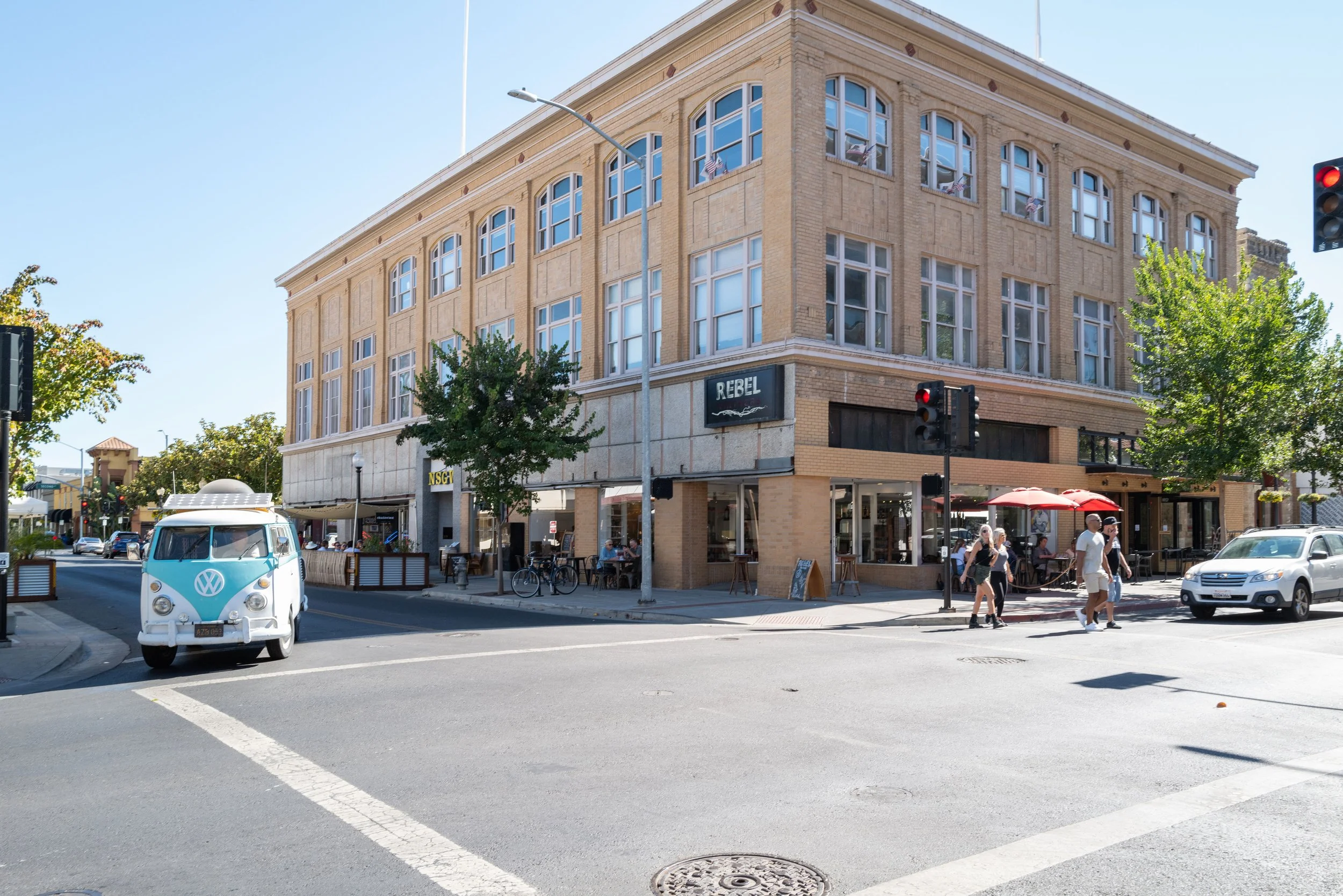 Corner street view with a brick building, outdoor dining area, pedestrians, a vintage Volkswagen van, and traffic lights.