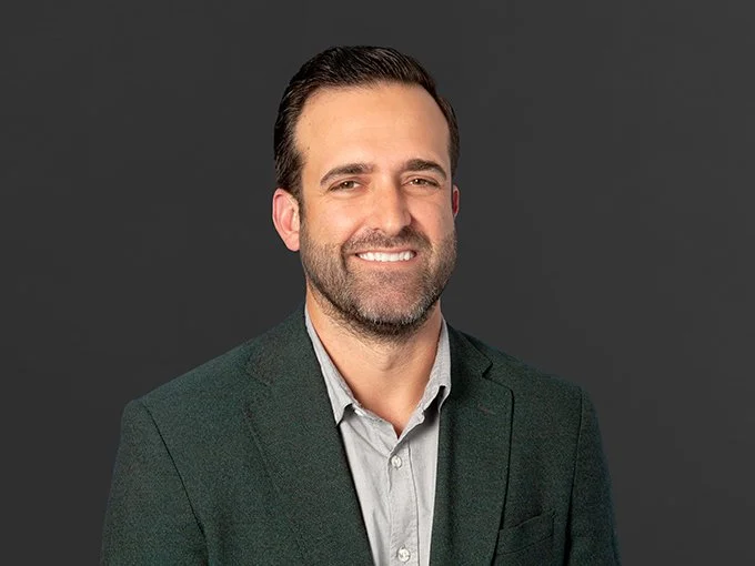 Man with dark hair and beard, wearing a dark blazer and light shirt, smiling against a dark gray background.