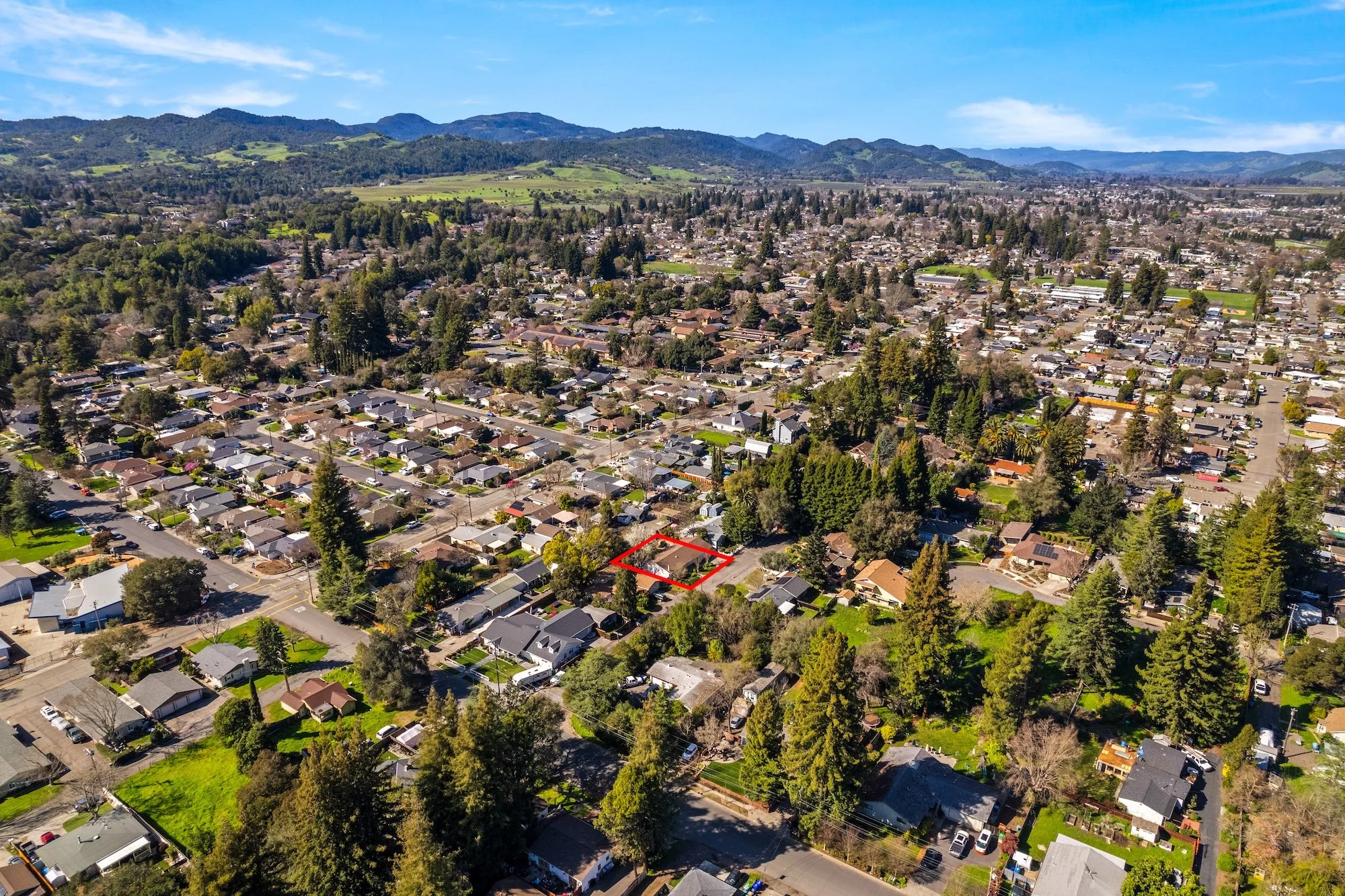 Aerial view of a suburban neighborhood with a specific property outlined in red, surrounded by trees and residential homes, with mountains and open land in the background under a blue sky.
