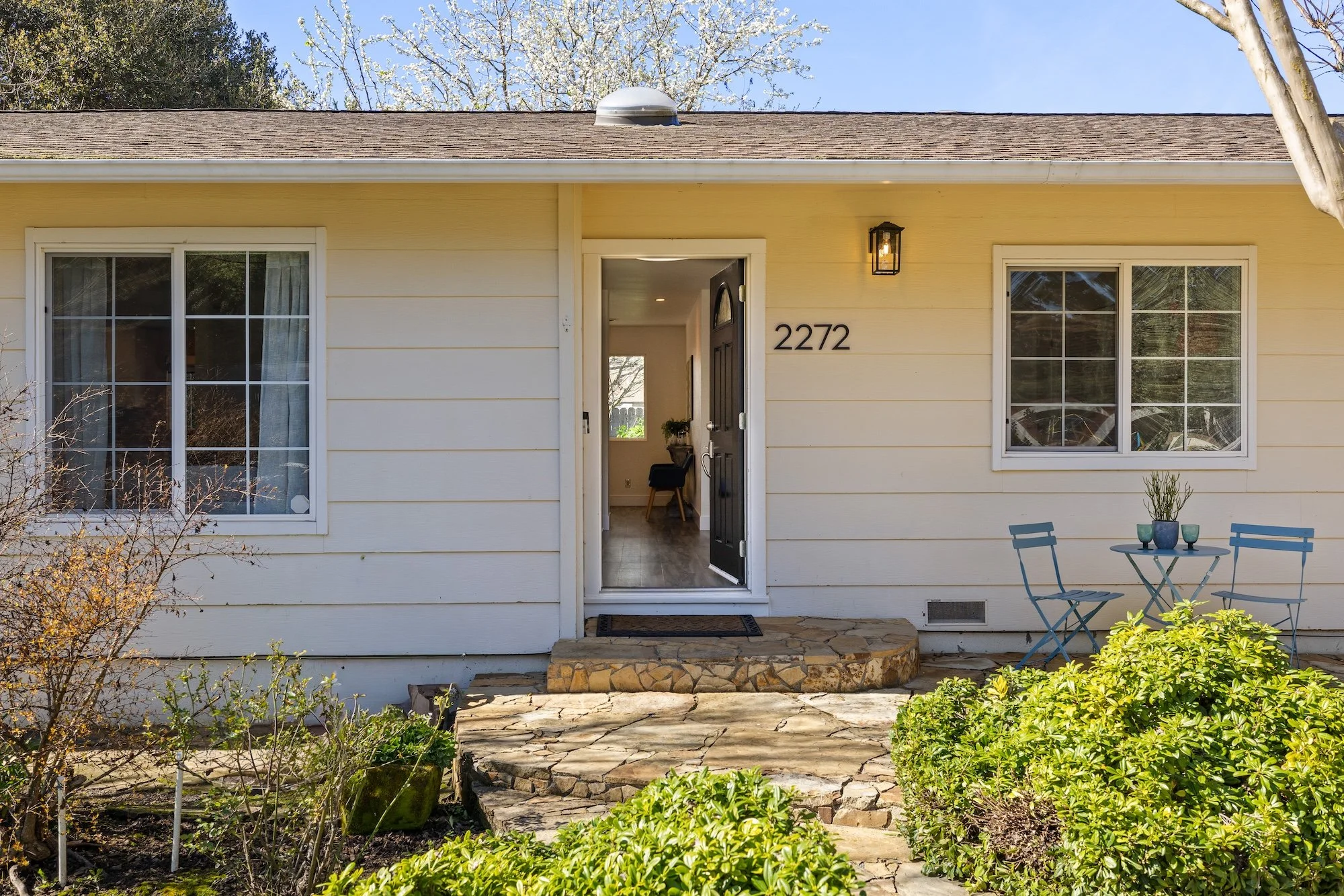 The front exterior of a house with beige siding, a stone step at the entrance, and a small patio with a table and two chairs. There are two large windows, a black front door, and a light fixture above the door. The house number '2272' is displayed ne