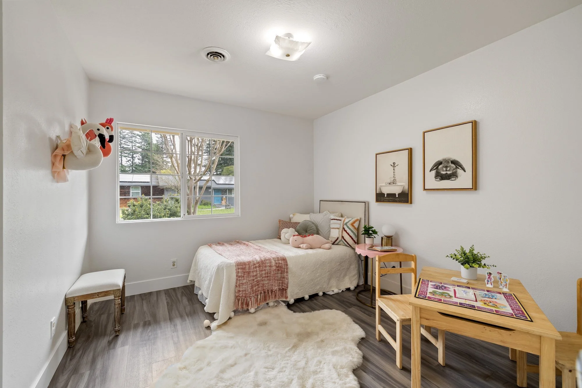 A bright bedroom with a single bed covered in white bedding and stuffed animals, a window showing trees outside, framed artwork on the wall, a small pink side table with a lamp and decor, a wooden table with a potted plant and magazines, a fluffy whi