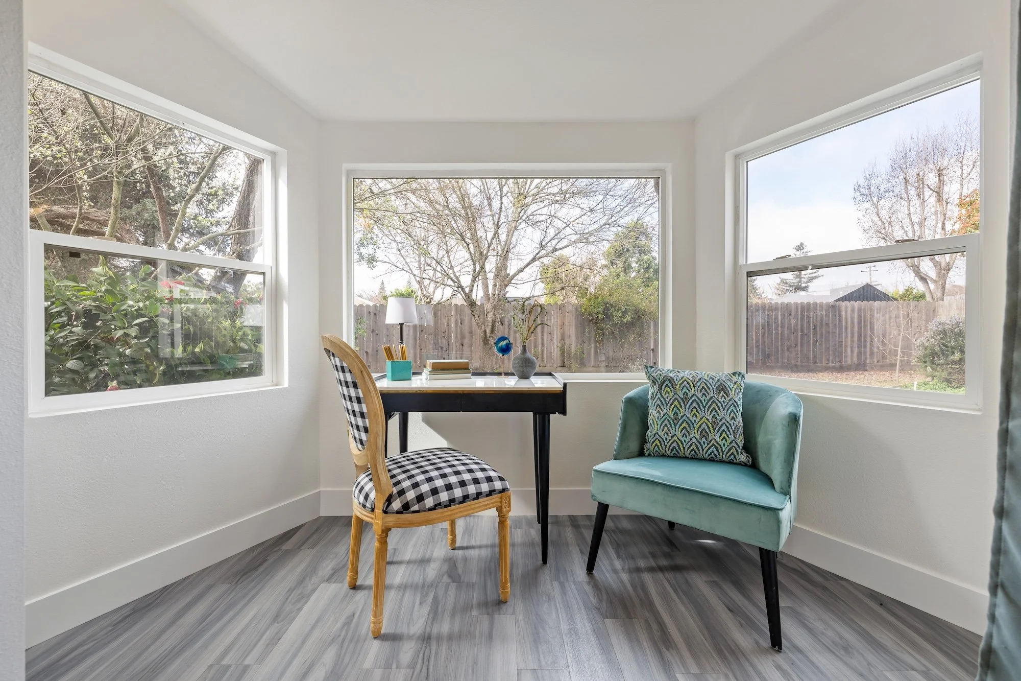 A bright sunroom with three large windows, white walls, a wooden desk with a chair and a small turquoise armchair with a patterned pillow, hardwood floor, and trees visible outside.