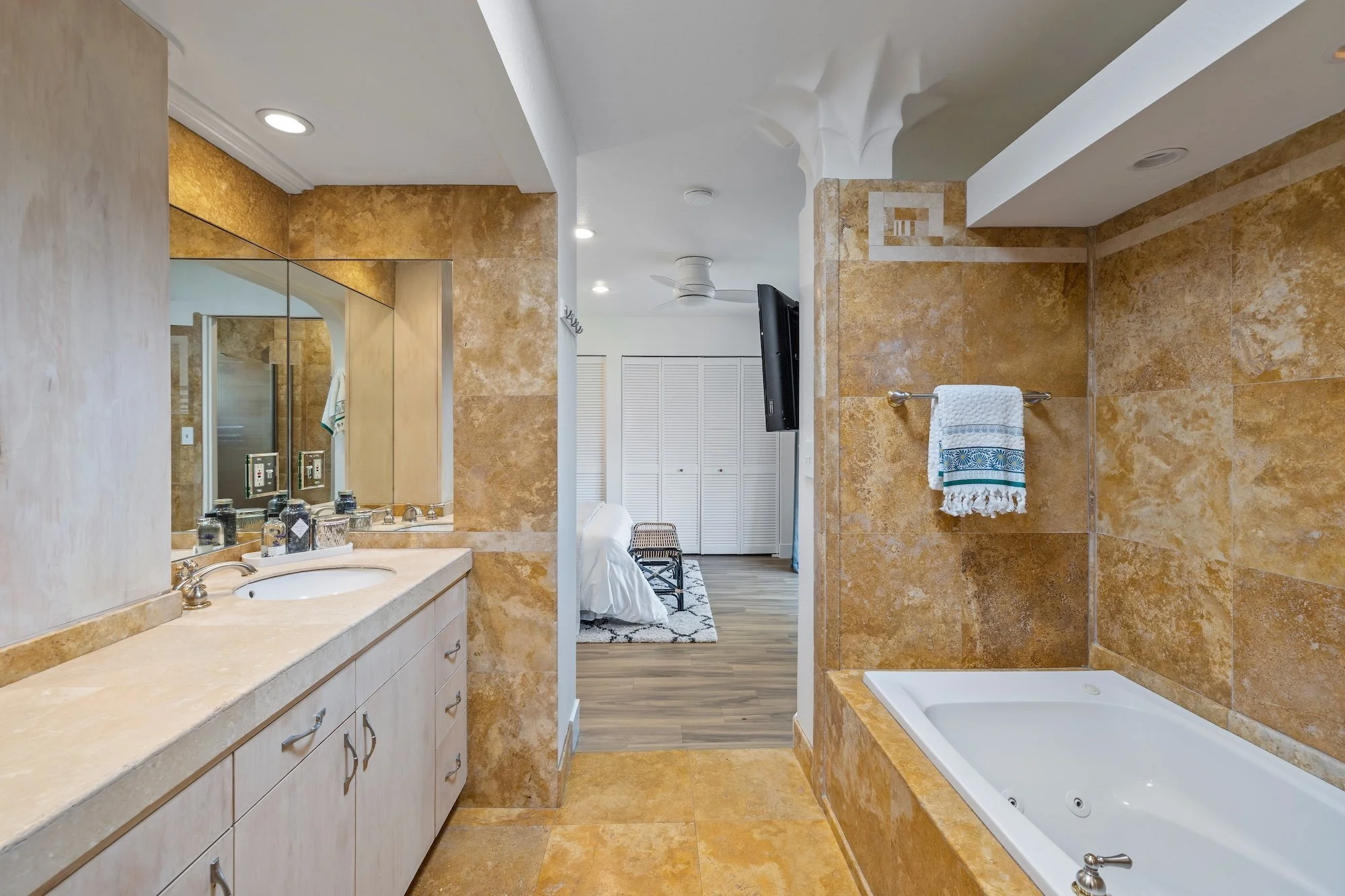 View of a bathroom with a beige vanity and sink on the left, large mirror above, and cupboard below. On the right, there's a built-in bathtub with beige tiles on the wall and around it, with a towel hanging on a rod. In the background, a bedroom with