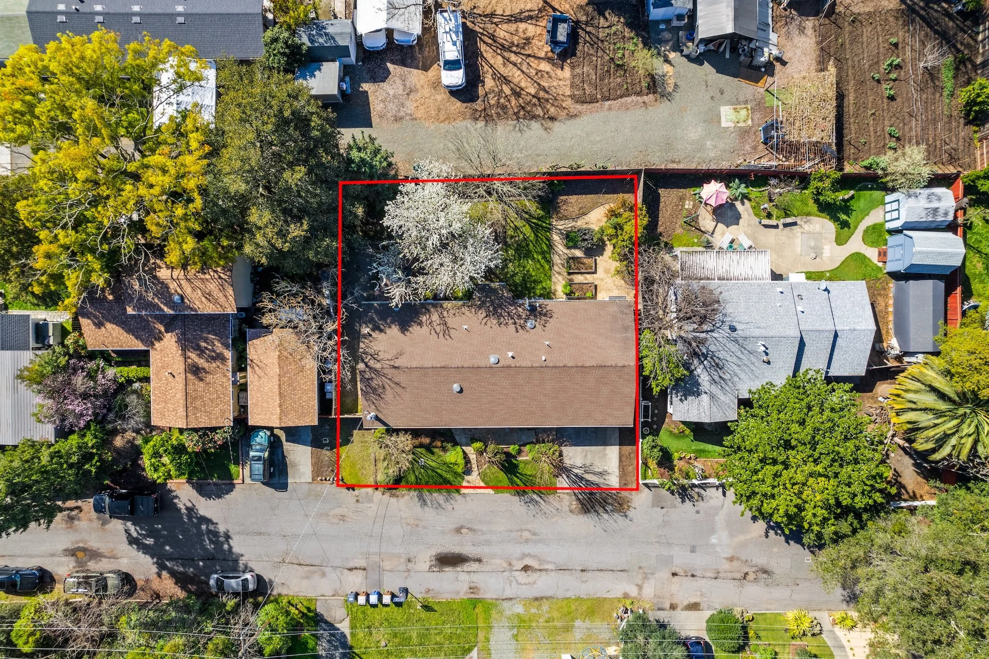 An aerial view of a residential backyard with a house in the center, surrounded by trees, a lawn, and neighboring houses and yards, with a red boundary lines marking the backyard.