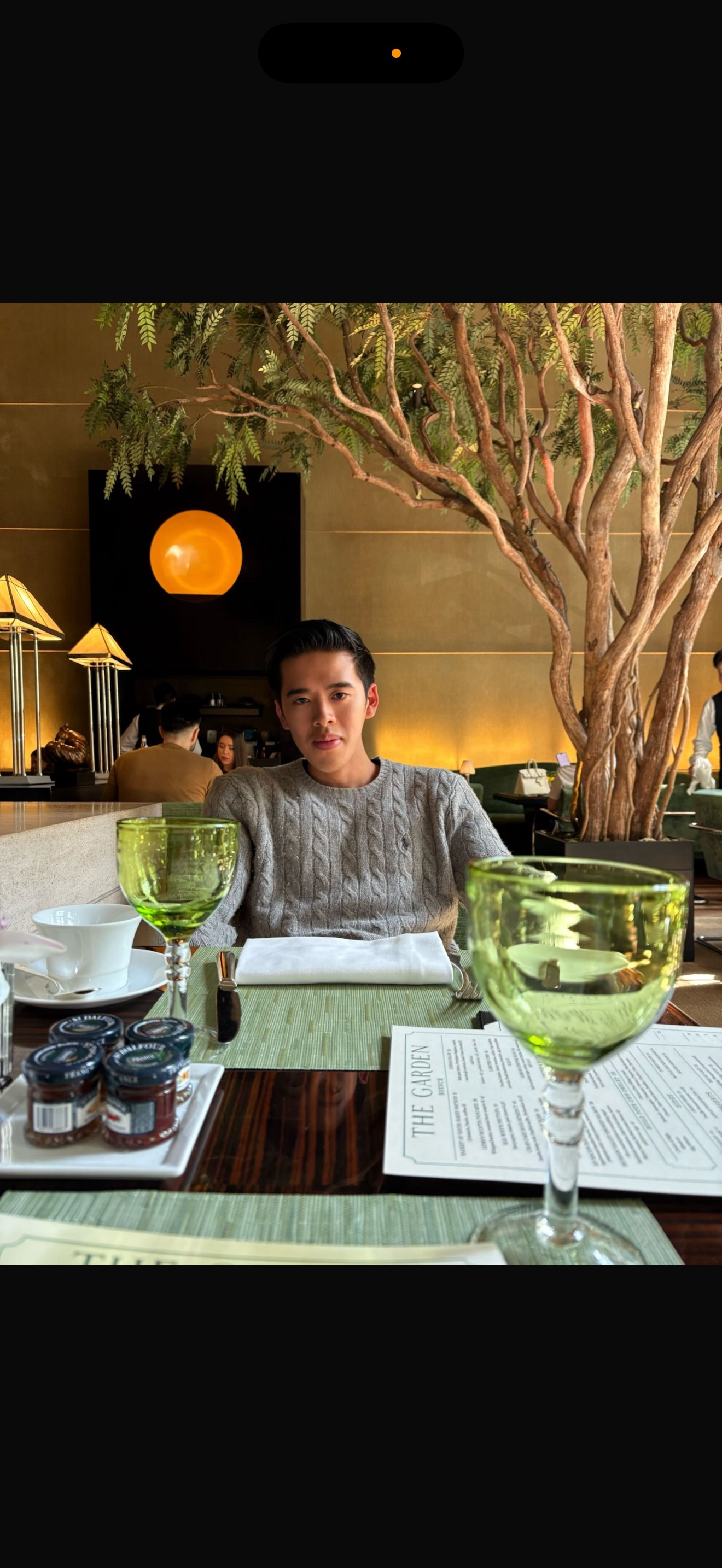 A young man with dark hair sitting at a restaurant table with a green glass, paper menu, and jars of jam, in a warmly lit dining area with a large tree and artistic wall decor in the background.