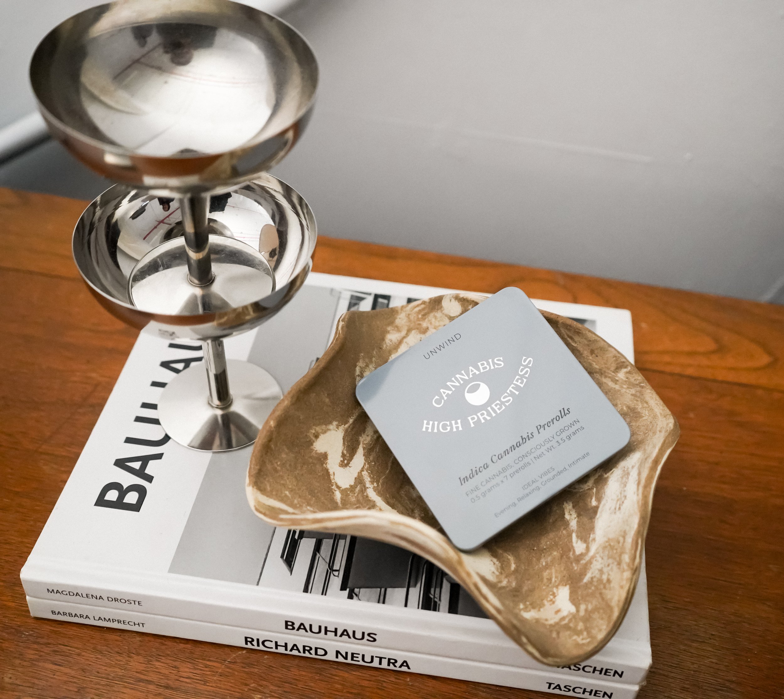 Close-up of a metallic sculpture with three shiny, conical shapes stacked vertically, placed on a table with a magazine underneath. The sculpture reflects the surrounding environment. There is a pamphlet or tag indicating the focus on cannabis and high potency, and a book or magazine about architectural design is partially visible underneath.