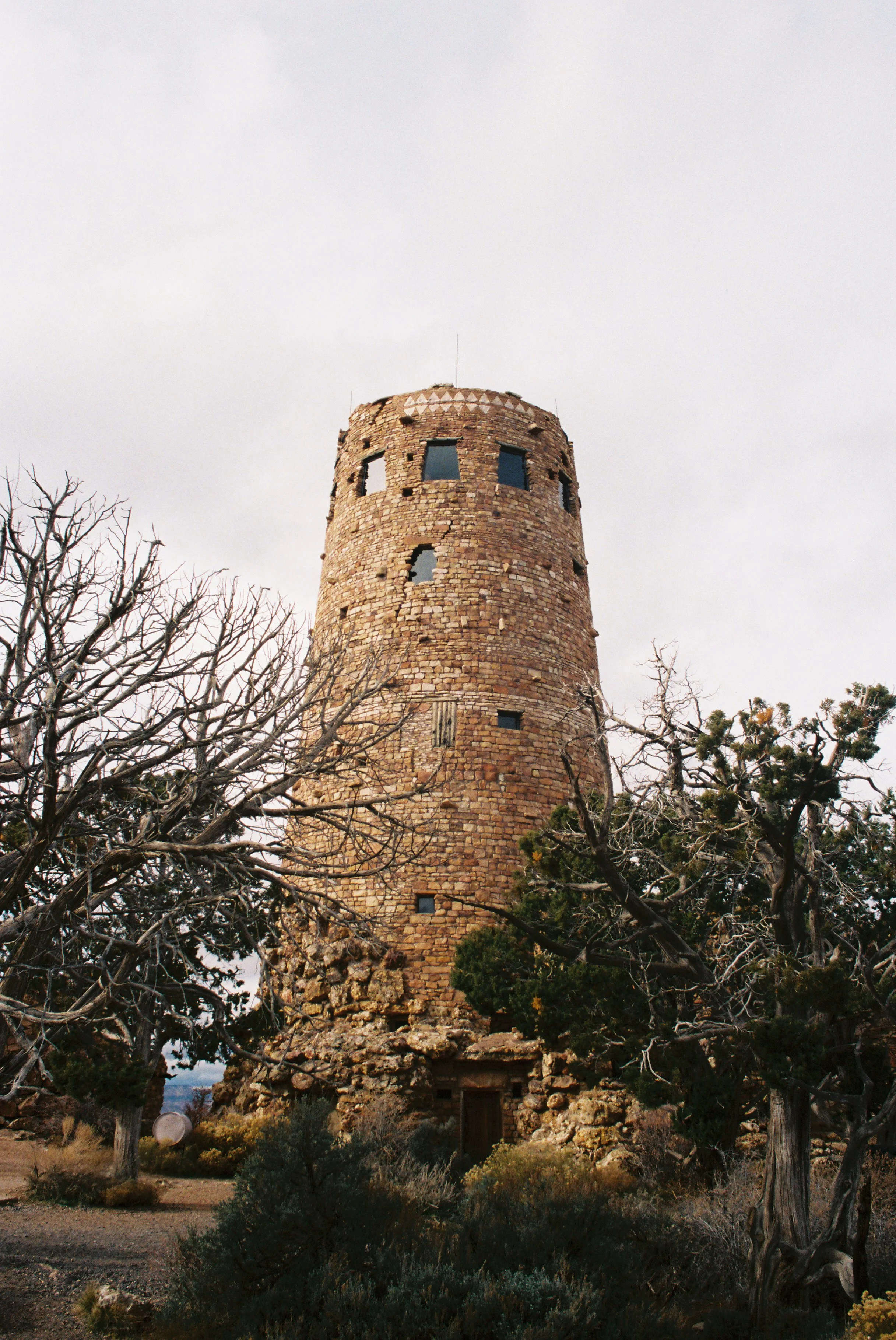 A tall, round stone tower with small windows, surrounded by leafless trees and shrubbery, under a cloudy sky.