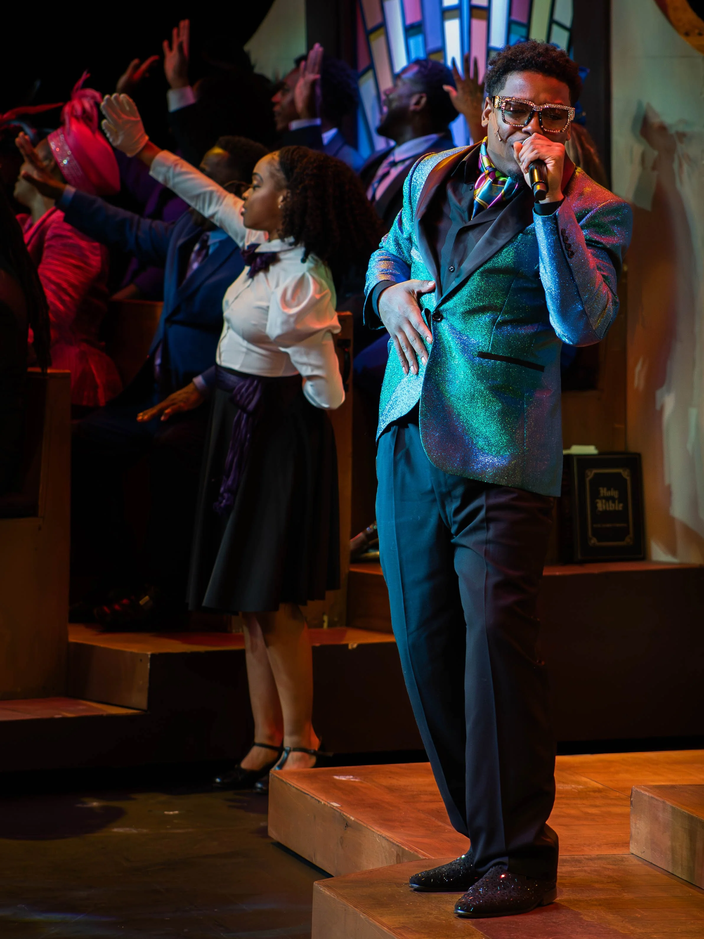 A man singing into a microphone on stage, wearing a shiny, multicolored blazer and glasses, with people in church choir standing behind him.