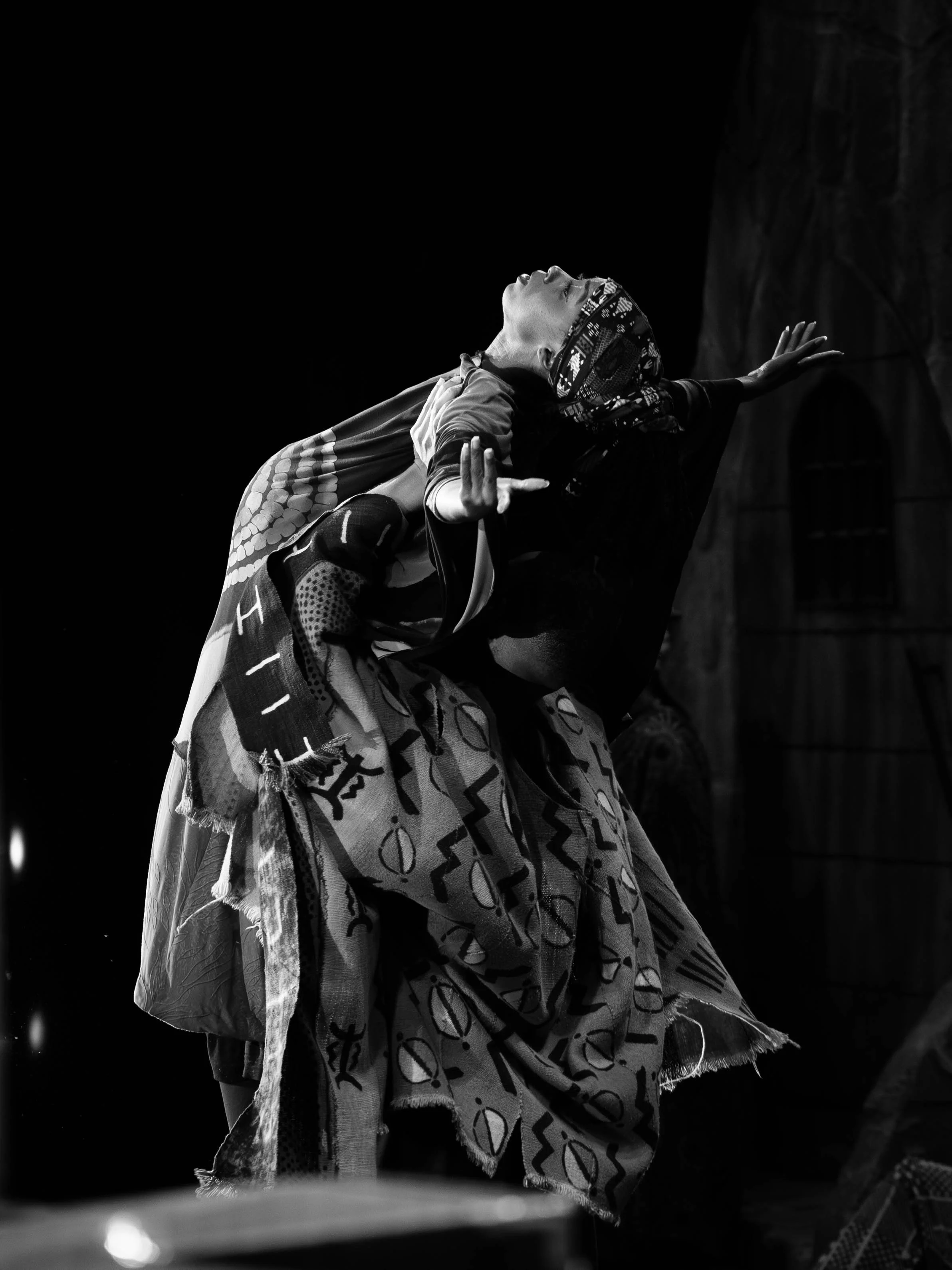 A person with a patterned headscarf and tasseled clothing is performing a dance or expressive gesture, with arms and head raised against a dark background.