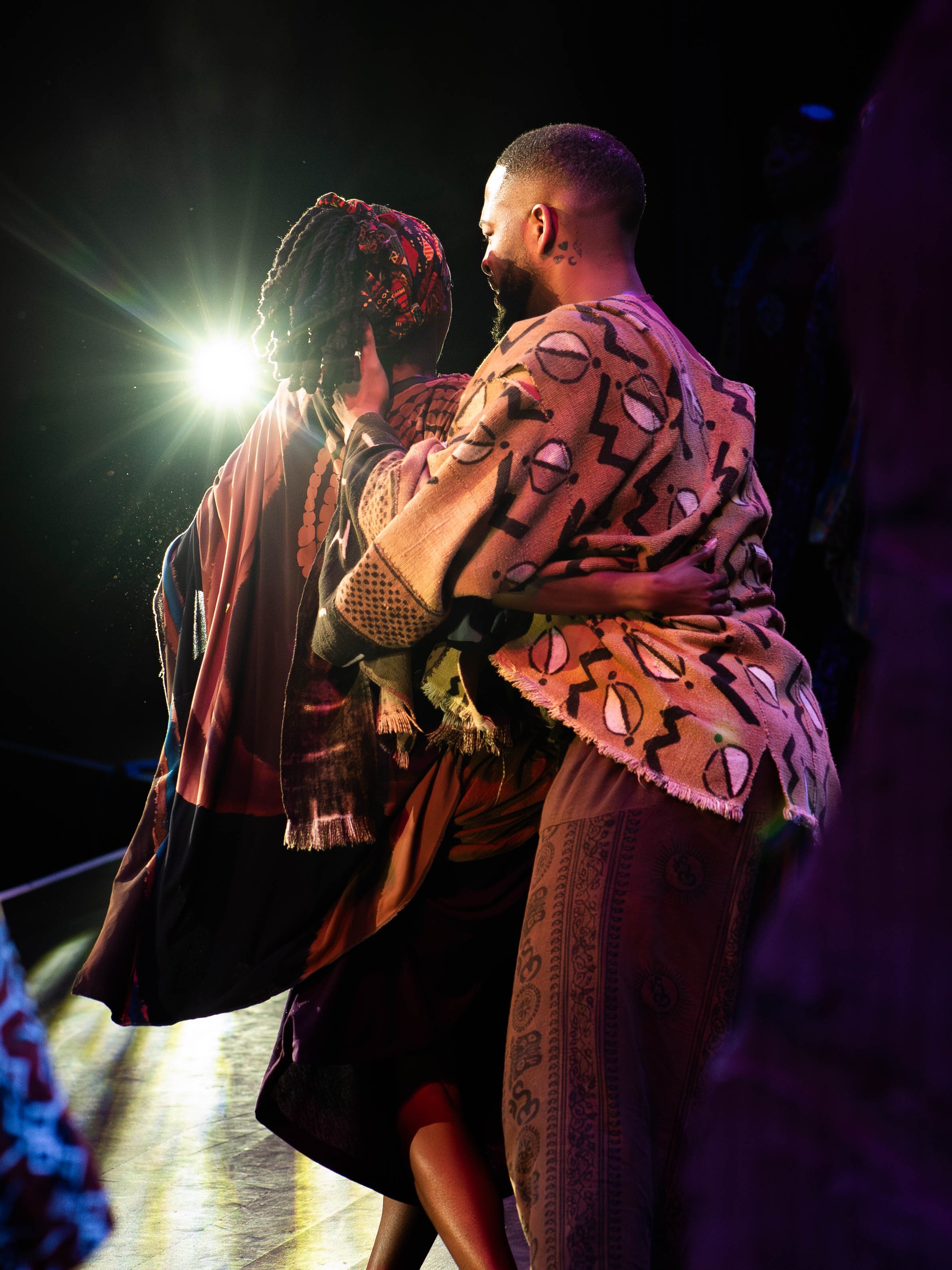 A couple dancing closely with bright backlighting, colorful patterned clothing, and a dark stage background.