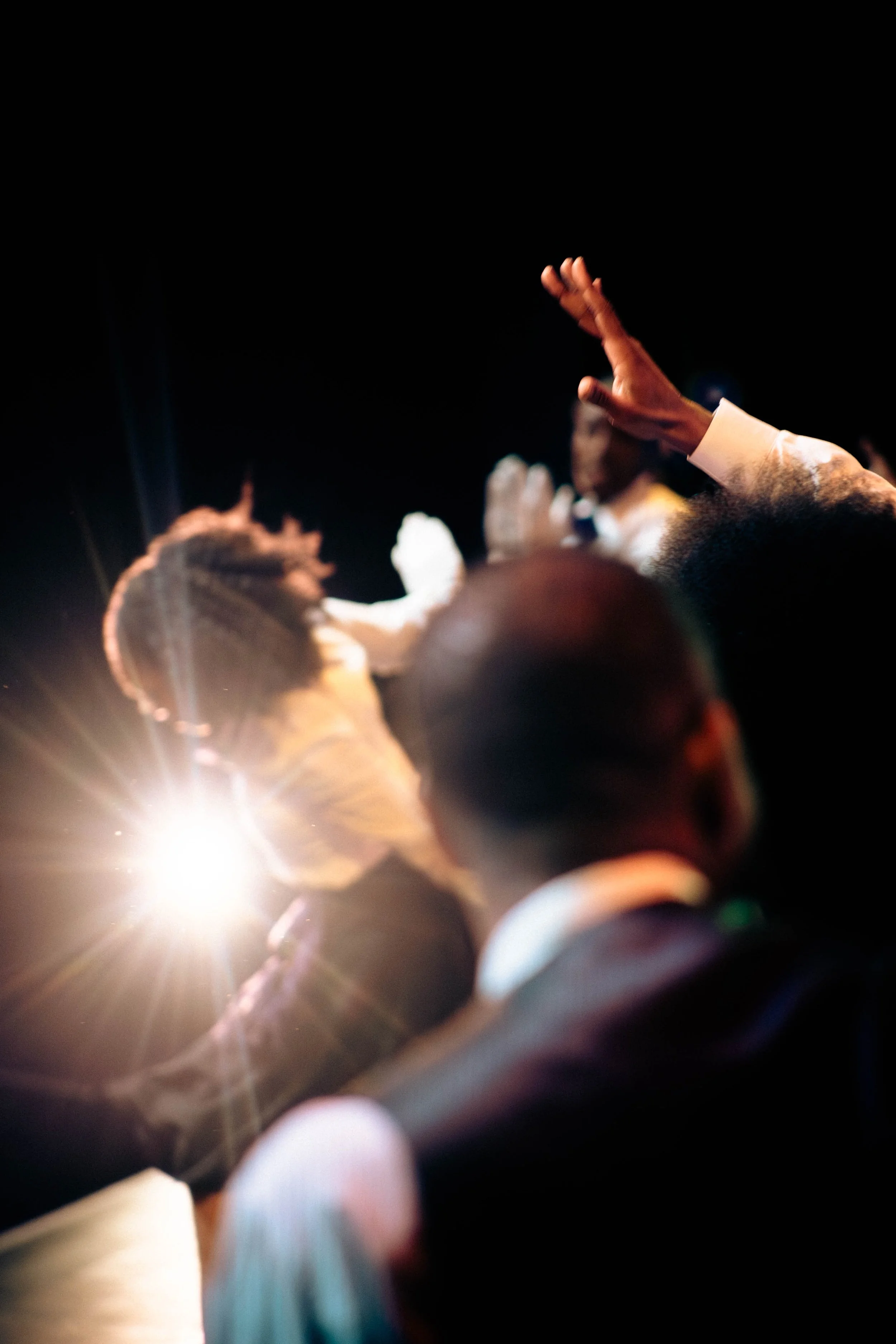 People attending a church service, with one person raising their hand and another person with braided hair, illuminated by bright stage lights.