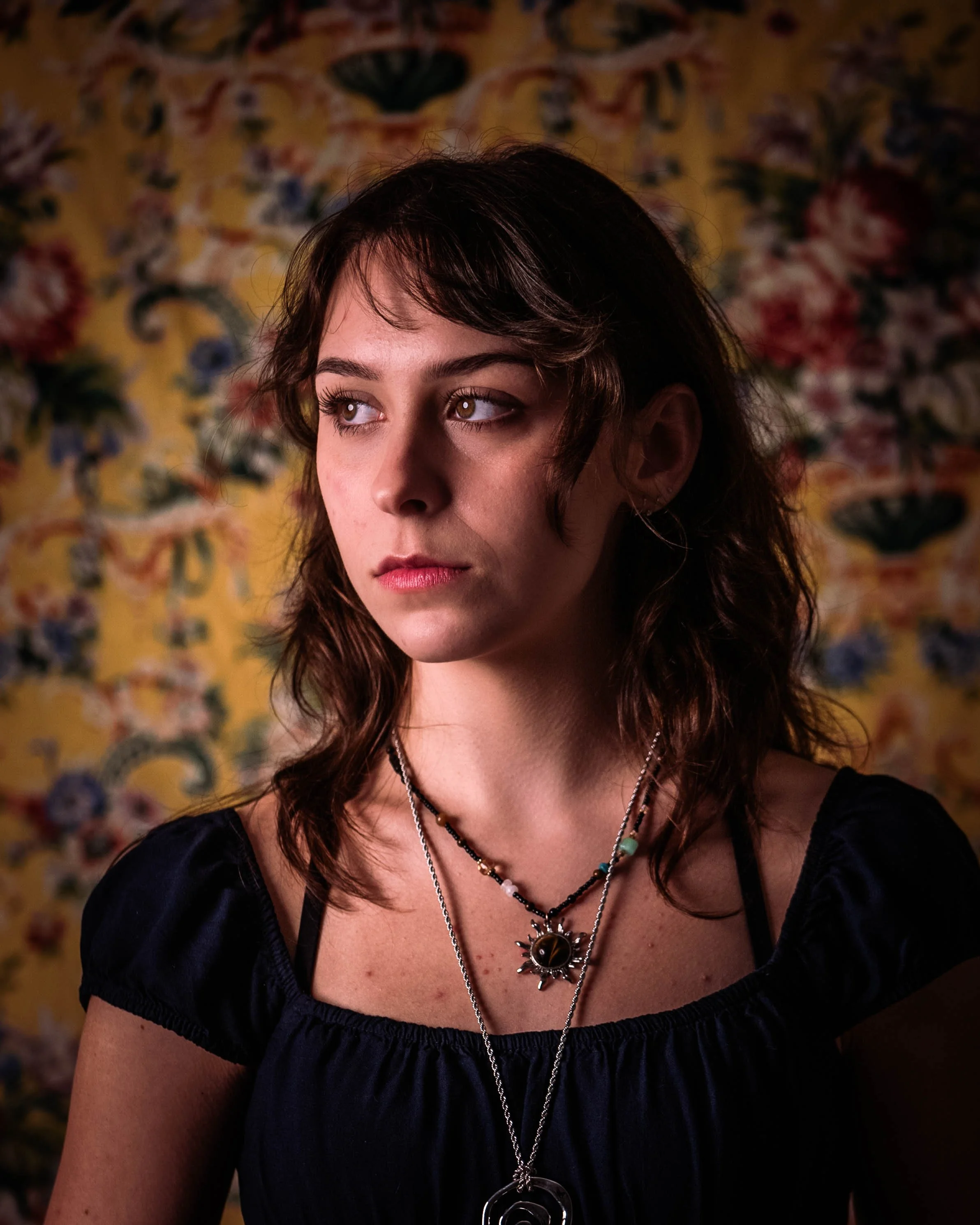 Close-up of a young woman with brown, wavy hair, wearing layered necklaces, wearing a dark top, with a colorful floral wallpaper background.