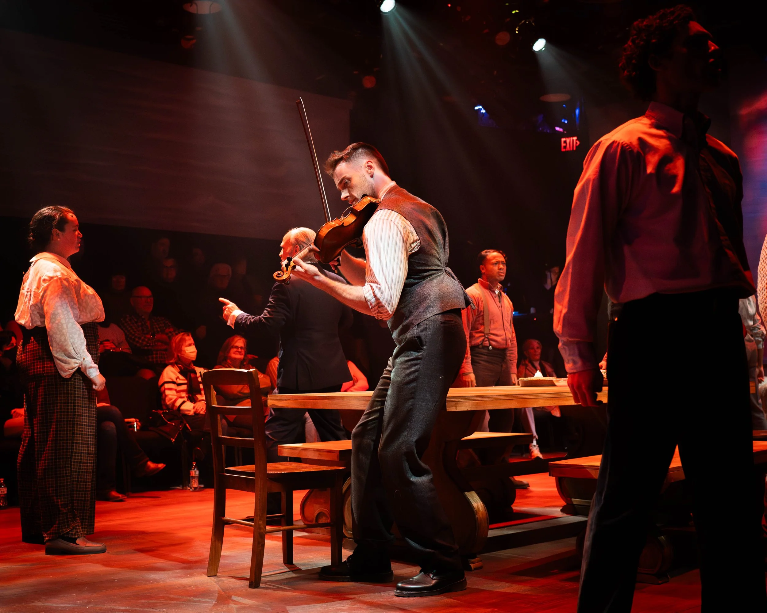 A violinist performing on stage with an audience watching, surrounded by other performers in vintage-style clothing.