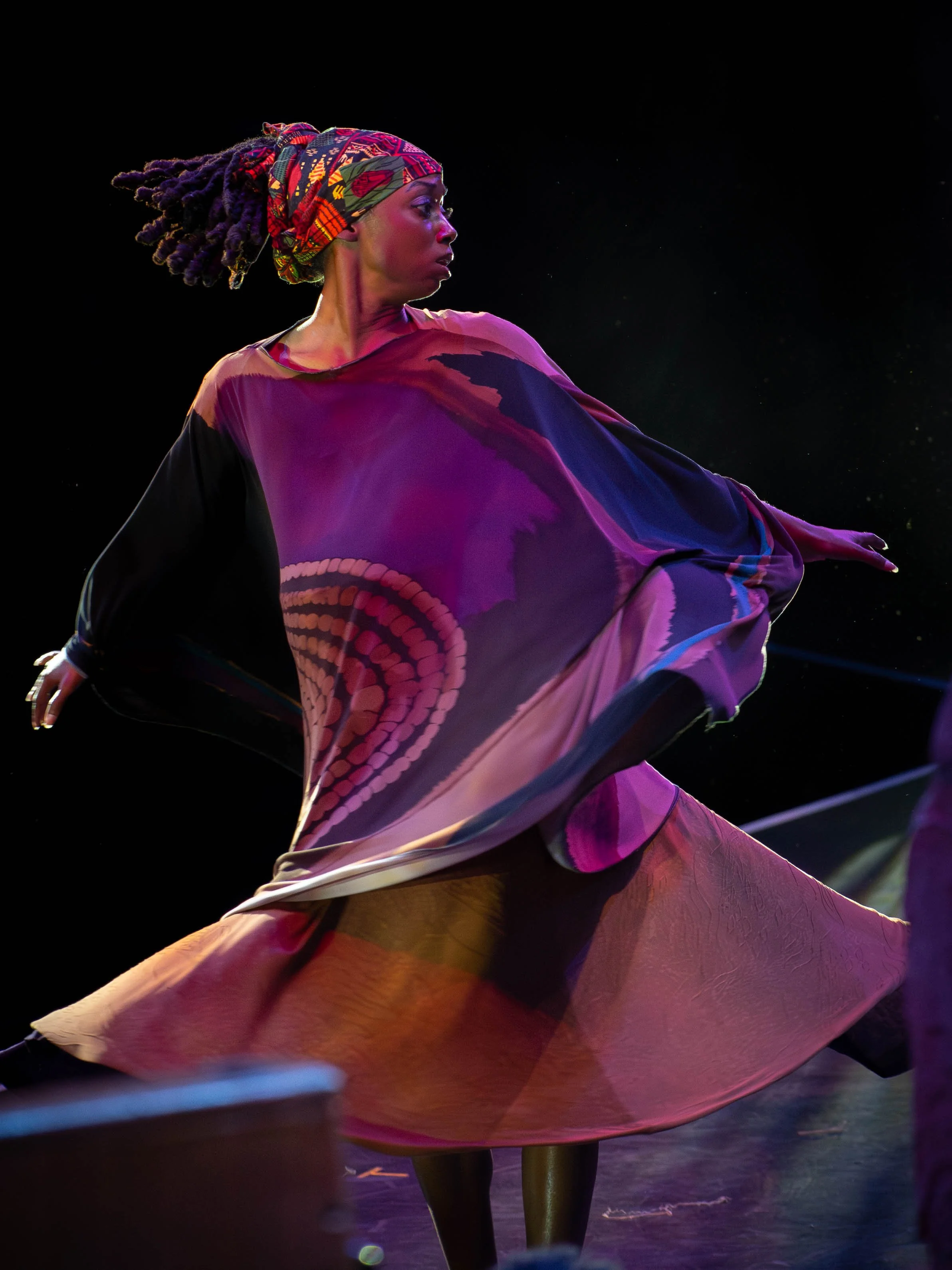 A woman with dreadlocks, wearing a colorful headscarf, performs on stage wearing a flowing multicolored dress with purple, pink, and black hues.