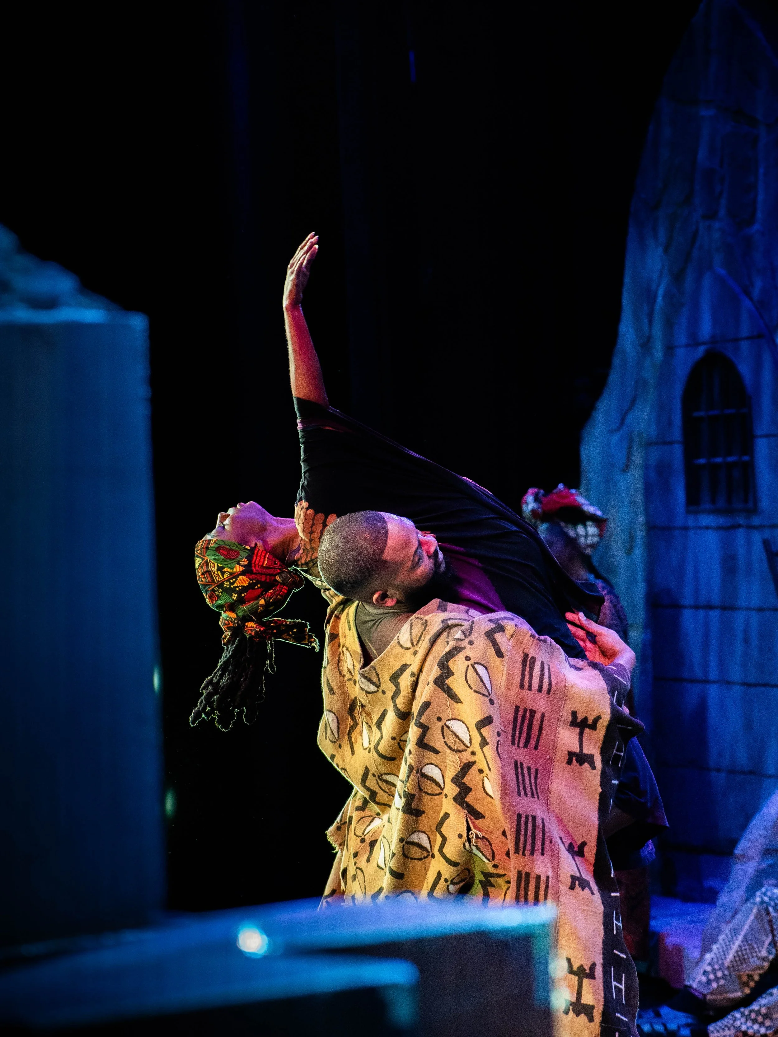 Two dancers perform a dance on stage, with one supporting the other who is leaning back with an arm raised, dressed in traditional African-inspired clothing and headwraps.