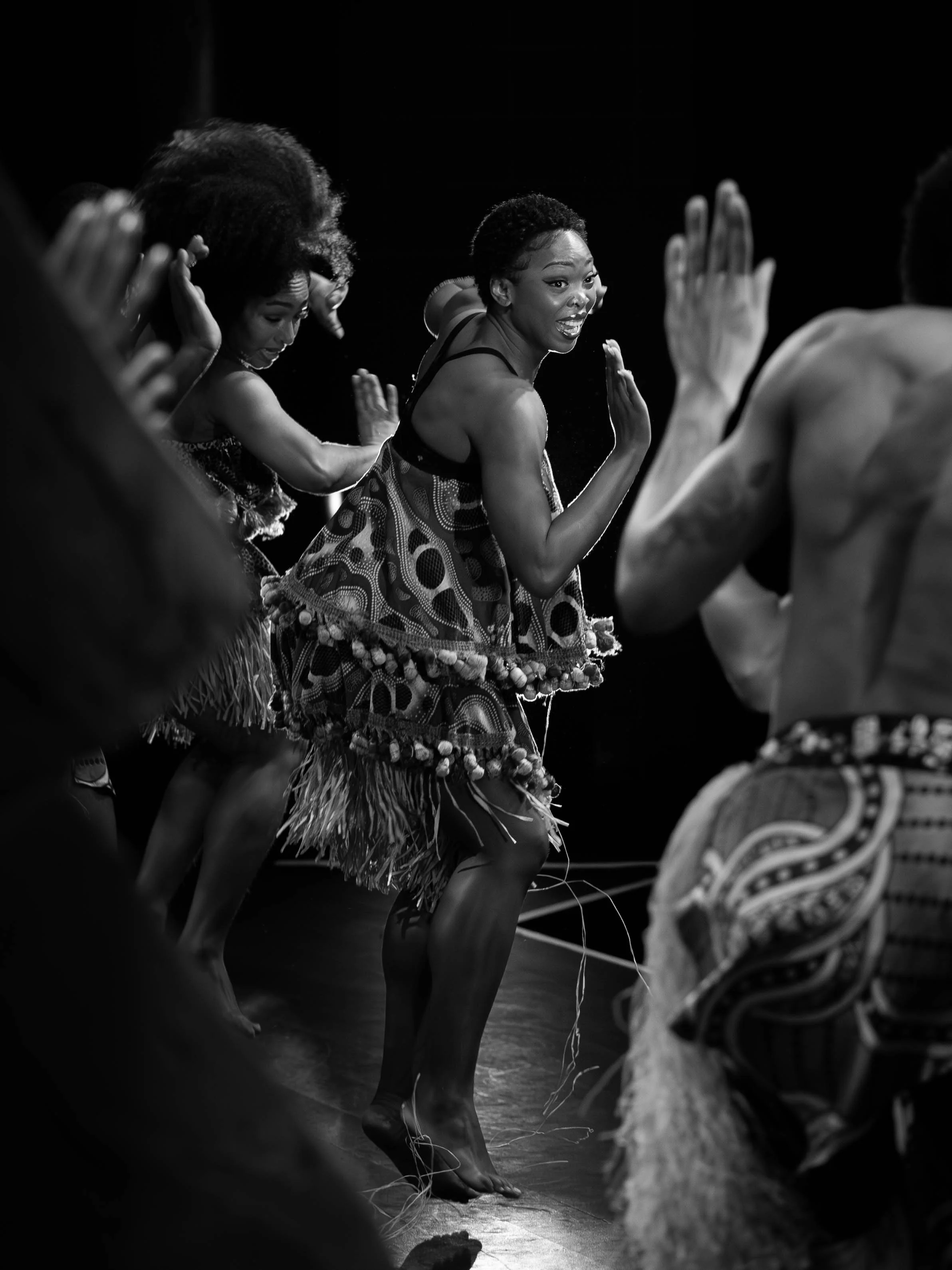 Group of dancers performing traditional Polynesian dance, wearing patterned skirts and barefoot, on stage with focused expressions.