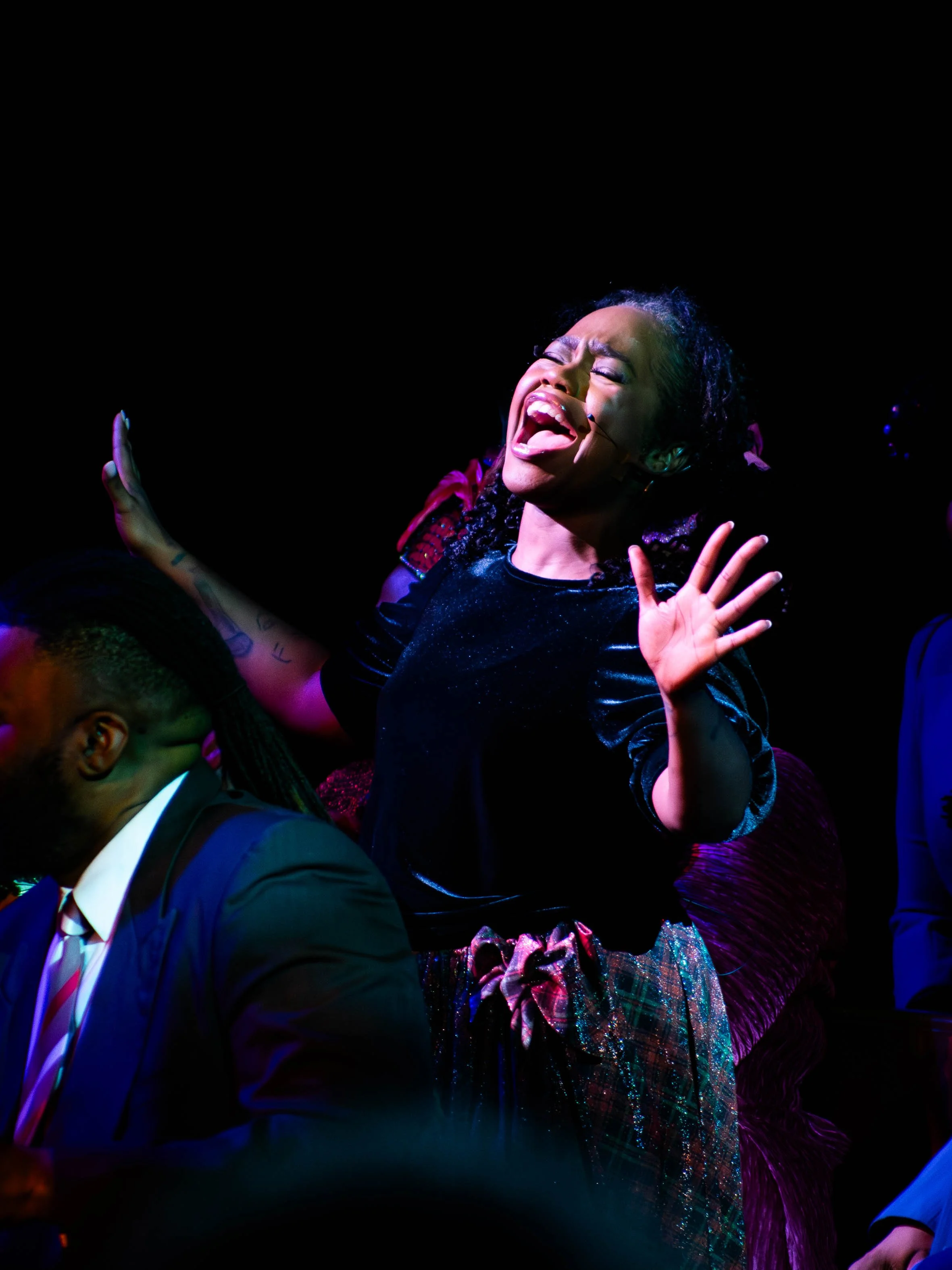 A woman singing passionately on stage with her eyes closed and hands raised, surrounded by other people in a dark environment.
