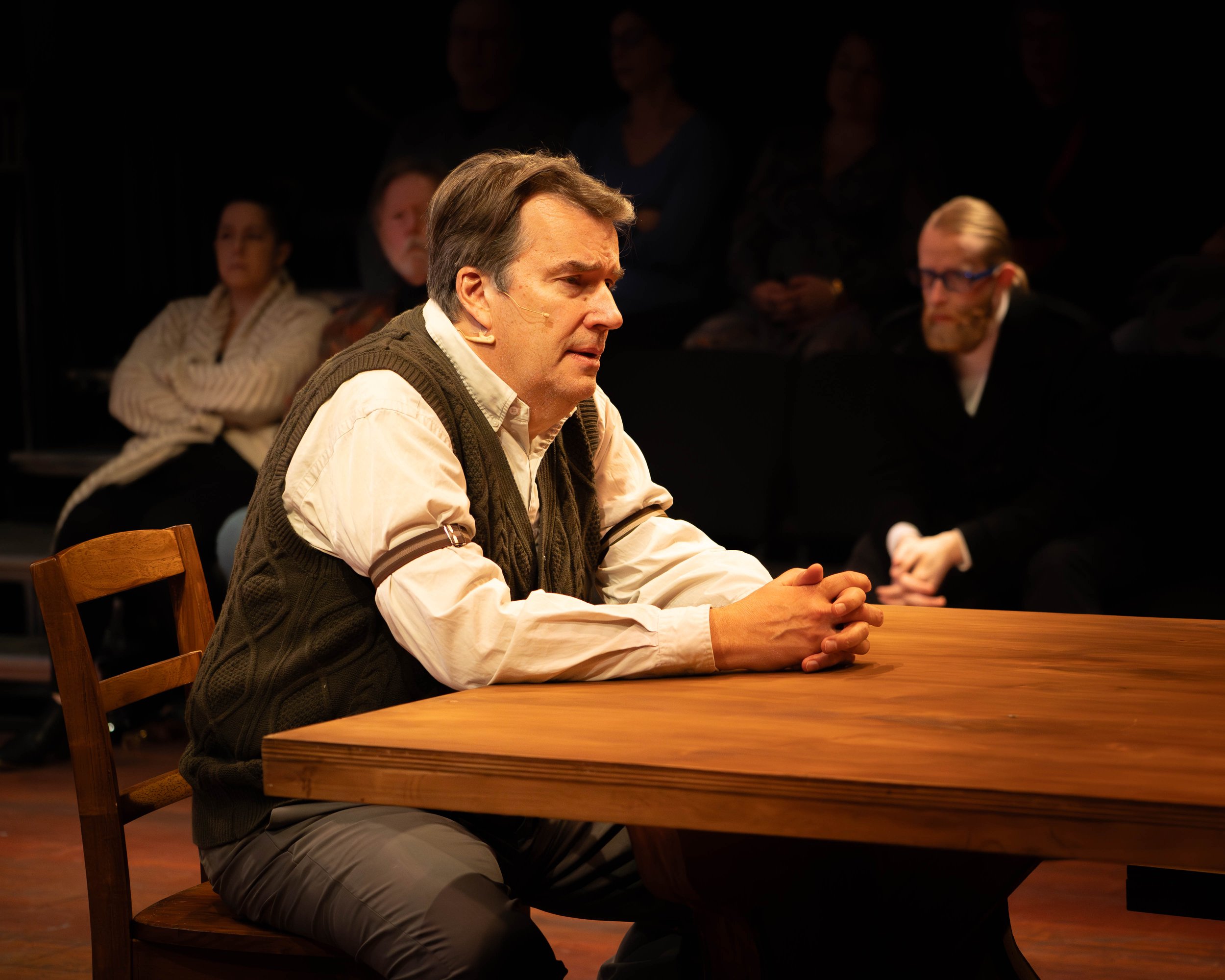A man with brown hair, wearing a white shirt and a brown vest, sitting at a wooden table with hands clasped, appearing contemplative, during a theatrical performance with an audience in the background.