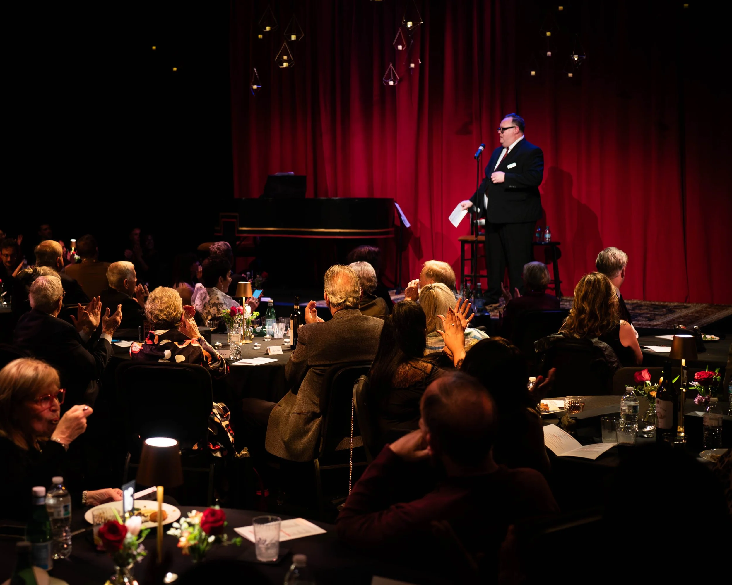 A man in a tuxedo and glasses speaking on stage at a formal event, with a red curtain backdrop and audience clapping at round tables with floral centerpieces.
