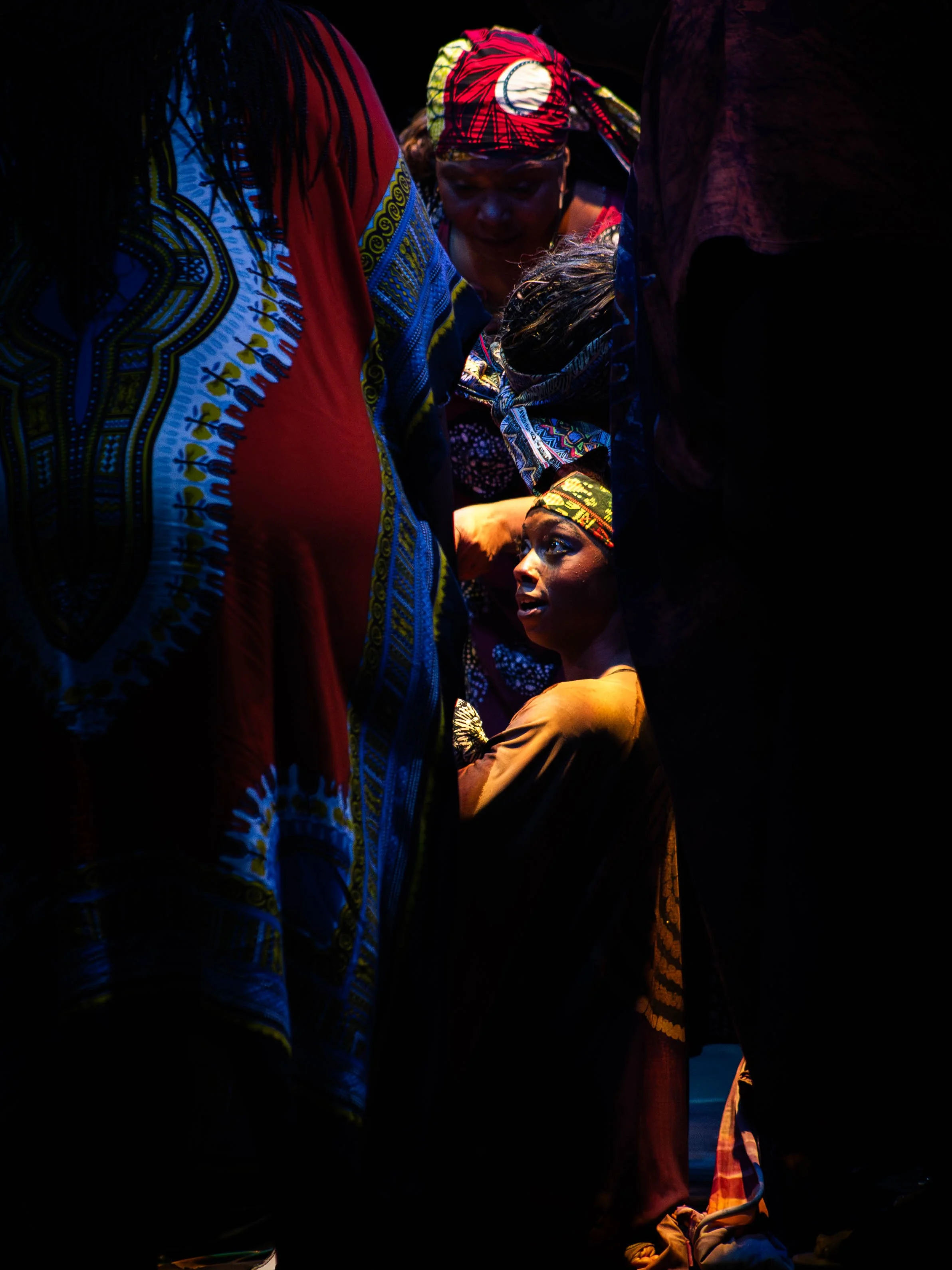 Group of people, mostly women, wearing colorful traditional African clothing and head wraps, gathered closely together in a dimly lit environment, with one woman sitting and looking up.
