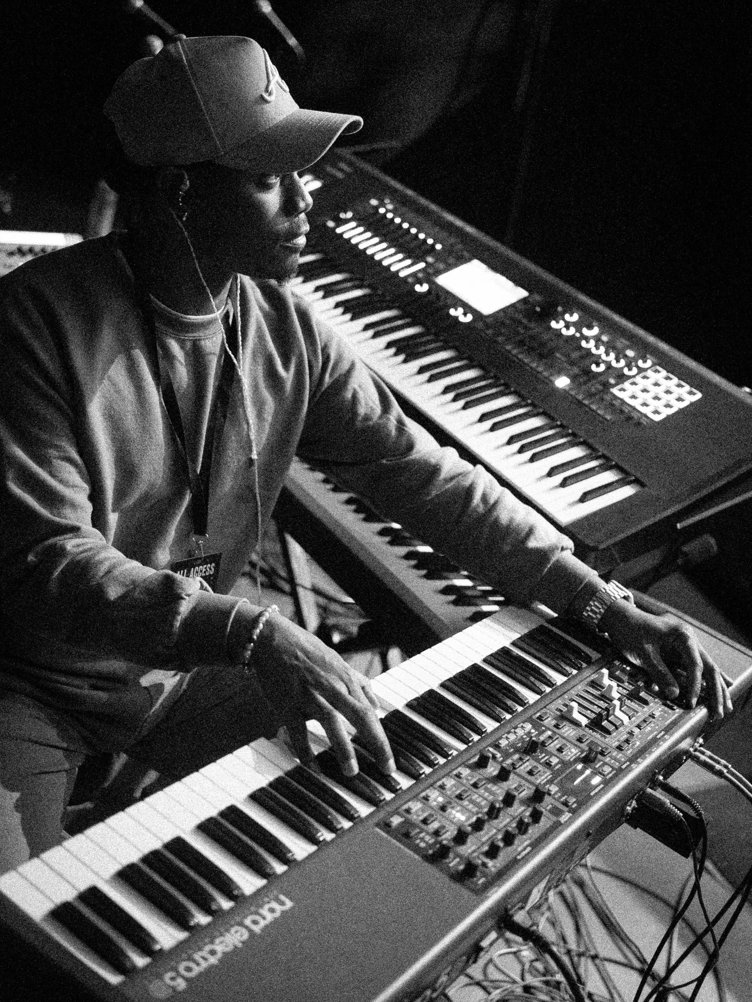 A person wearing a baseball cap and headphones playing a keyboard and a synthesizer in a recording studio.