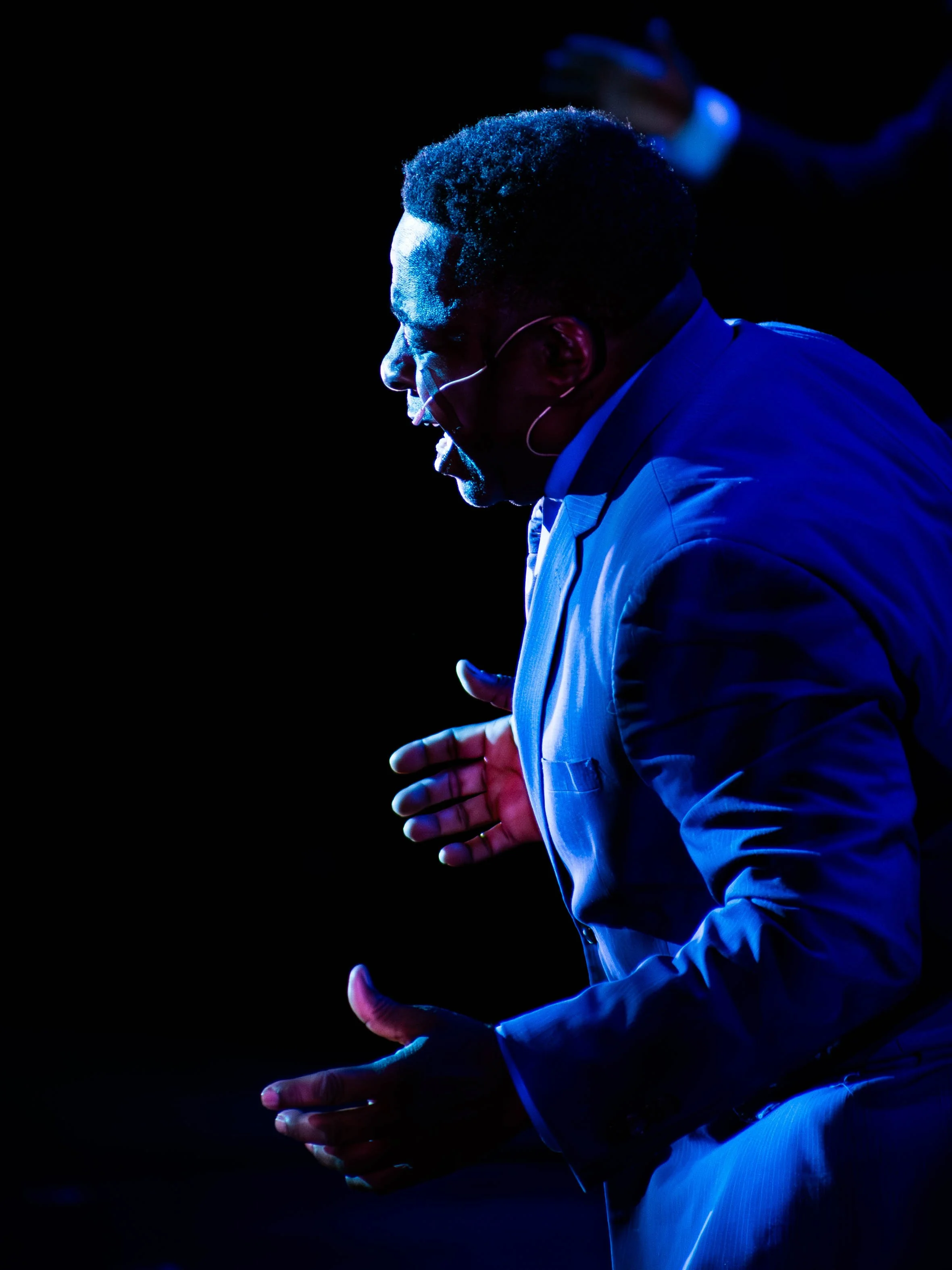 A man in a suit passionately speaking or singing on stage, illuminated by blue lighting, with his mouth open and hands gesturing.