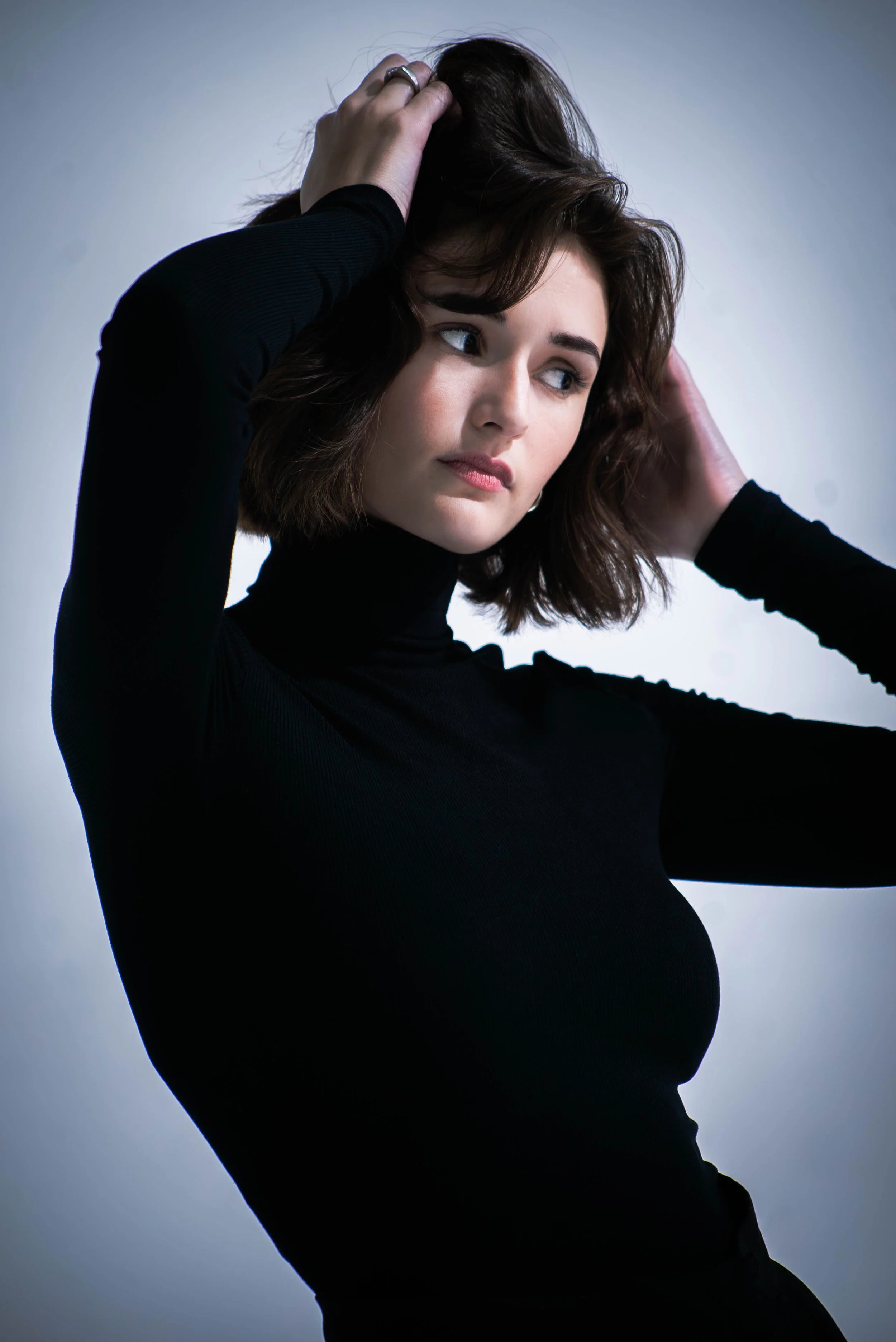 A woman with brown hair wearing a black turtleneck, posing against a gray background, with her hands in her hair.