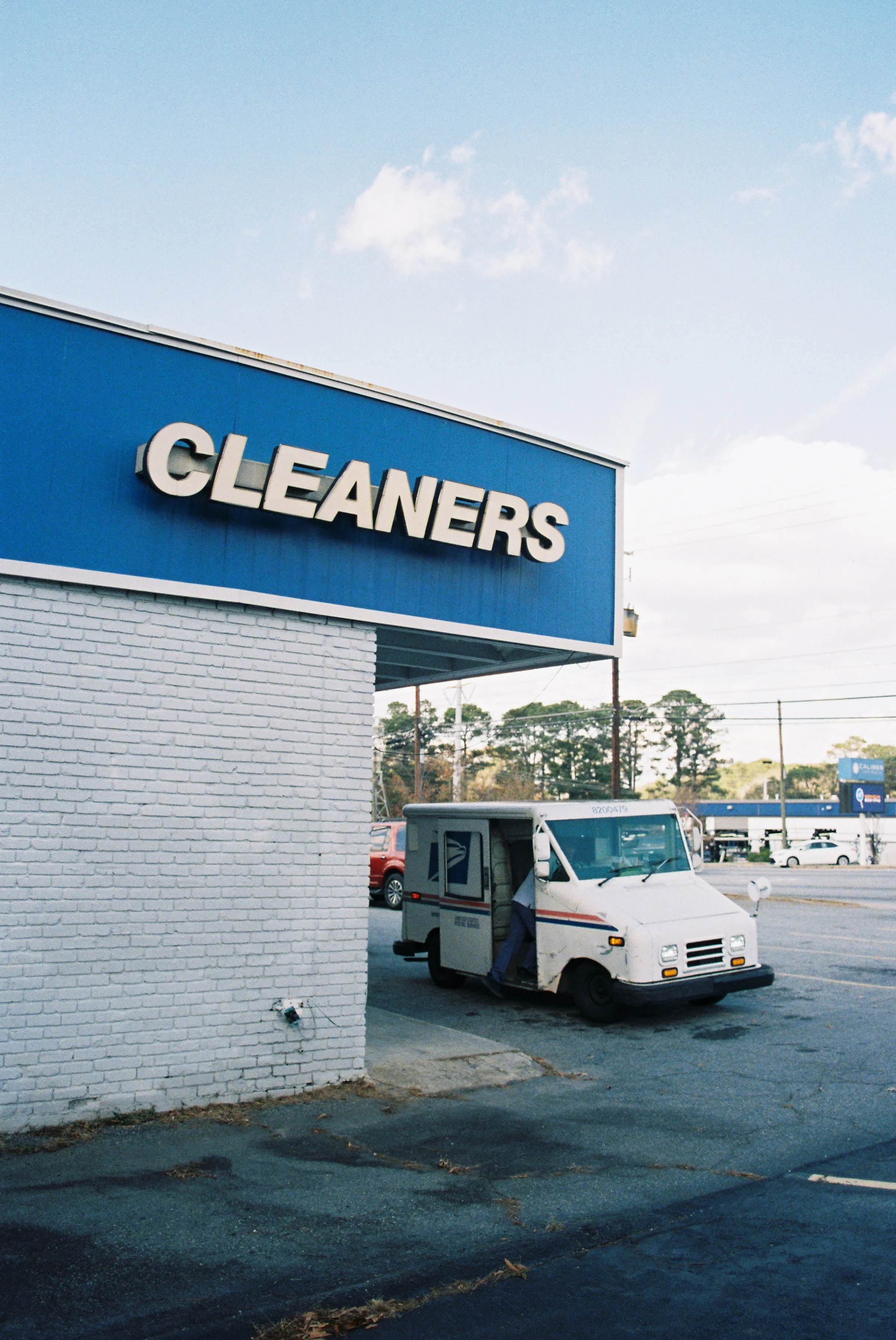 A blue-and-white cleaning supplies store with a prominent sign that says 'CLEANERS'. There is a small USPS mail delivery truck parked in front of the store, partially under the overhang, with open doors and a driver or worker partially visible inside