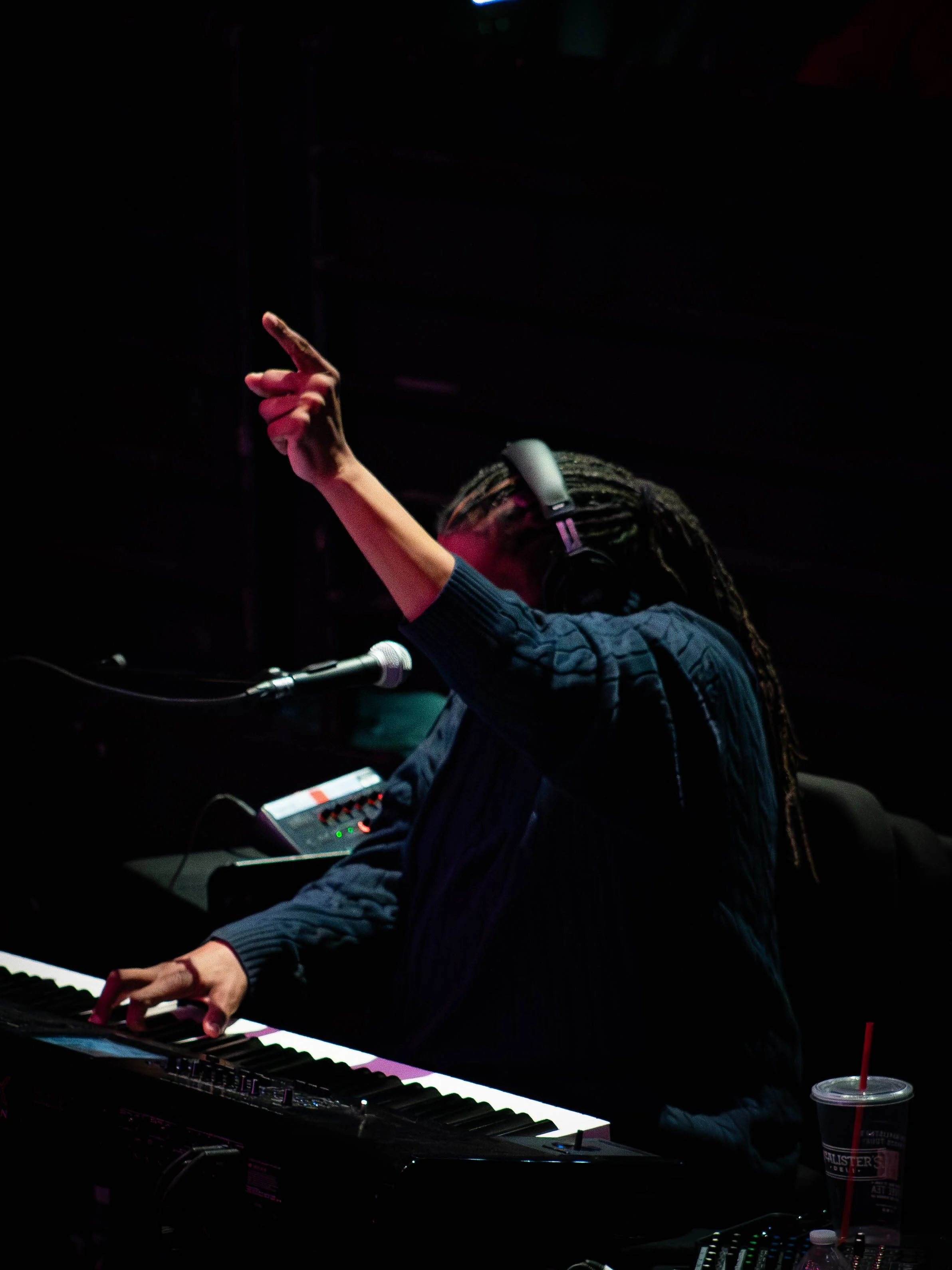 Person playing keyboard on stage, wearing headphones, holding a microphone, with a drink nearby, performing in a dark setting.