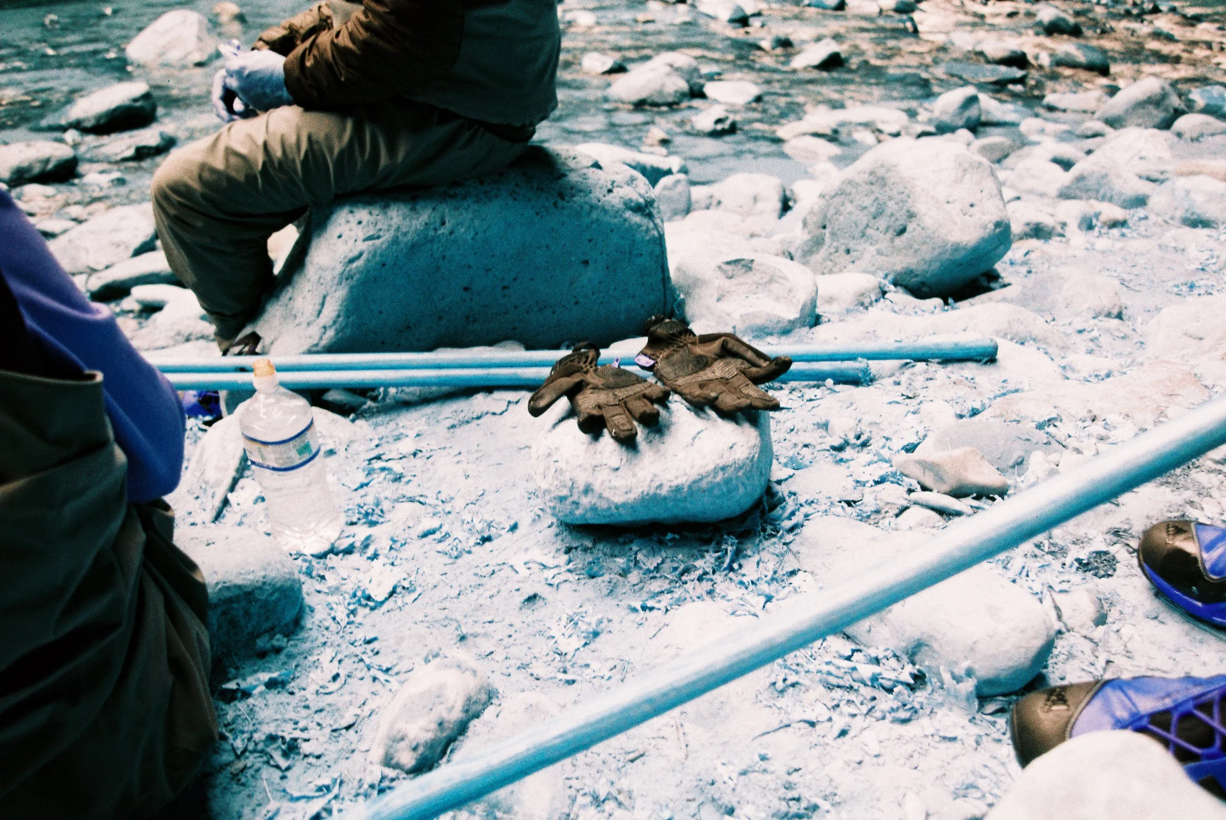 Gloves and a water bottle on the snow, with some poles and people sitting on rocks in a snowy outdoor area.