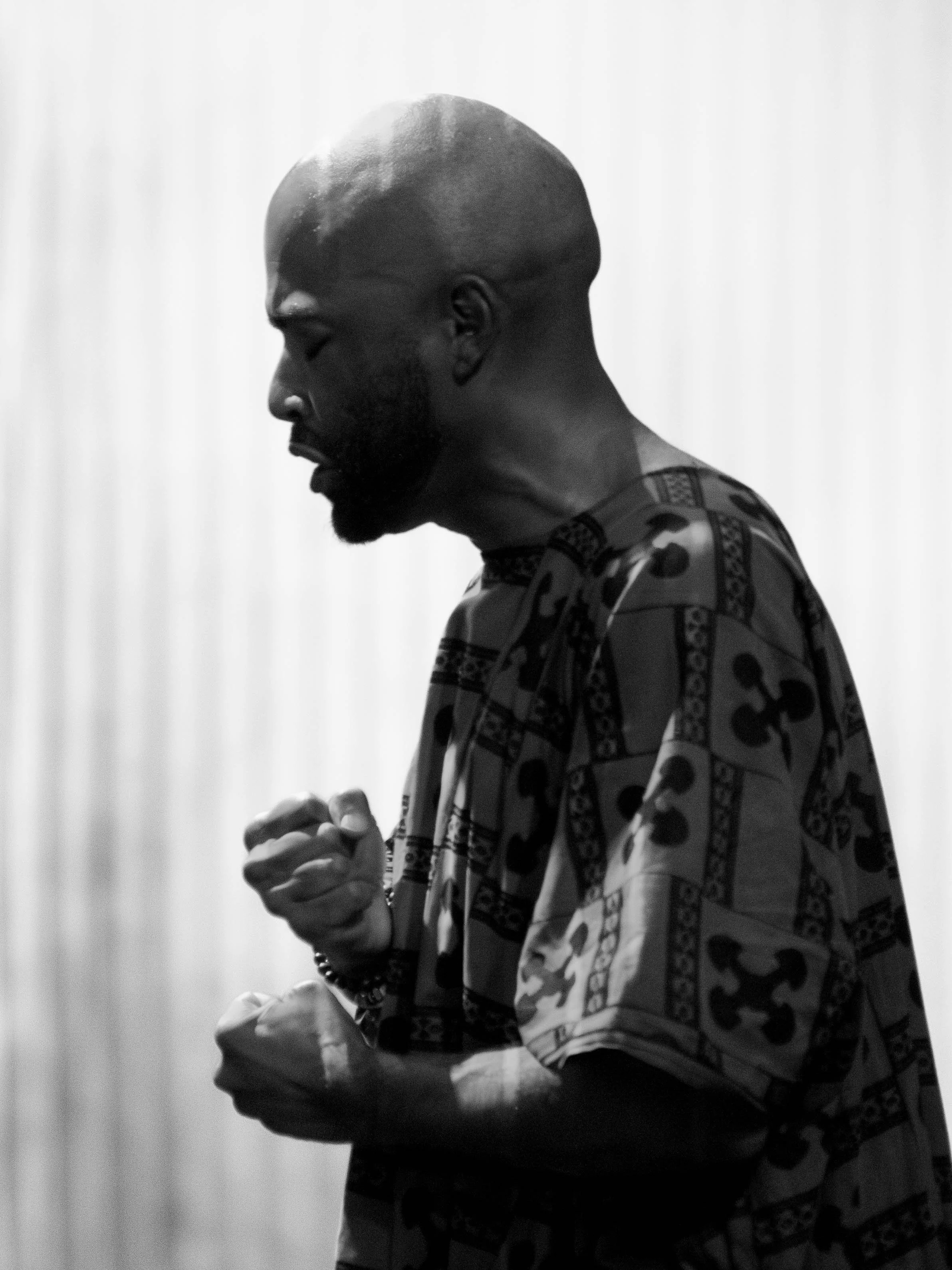 A man with a shaved head and beard, wearing a patterned shirt, has his eyes closed and hands clenched in front of him, in a moment of reflection or prayer.