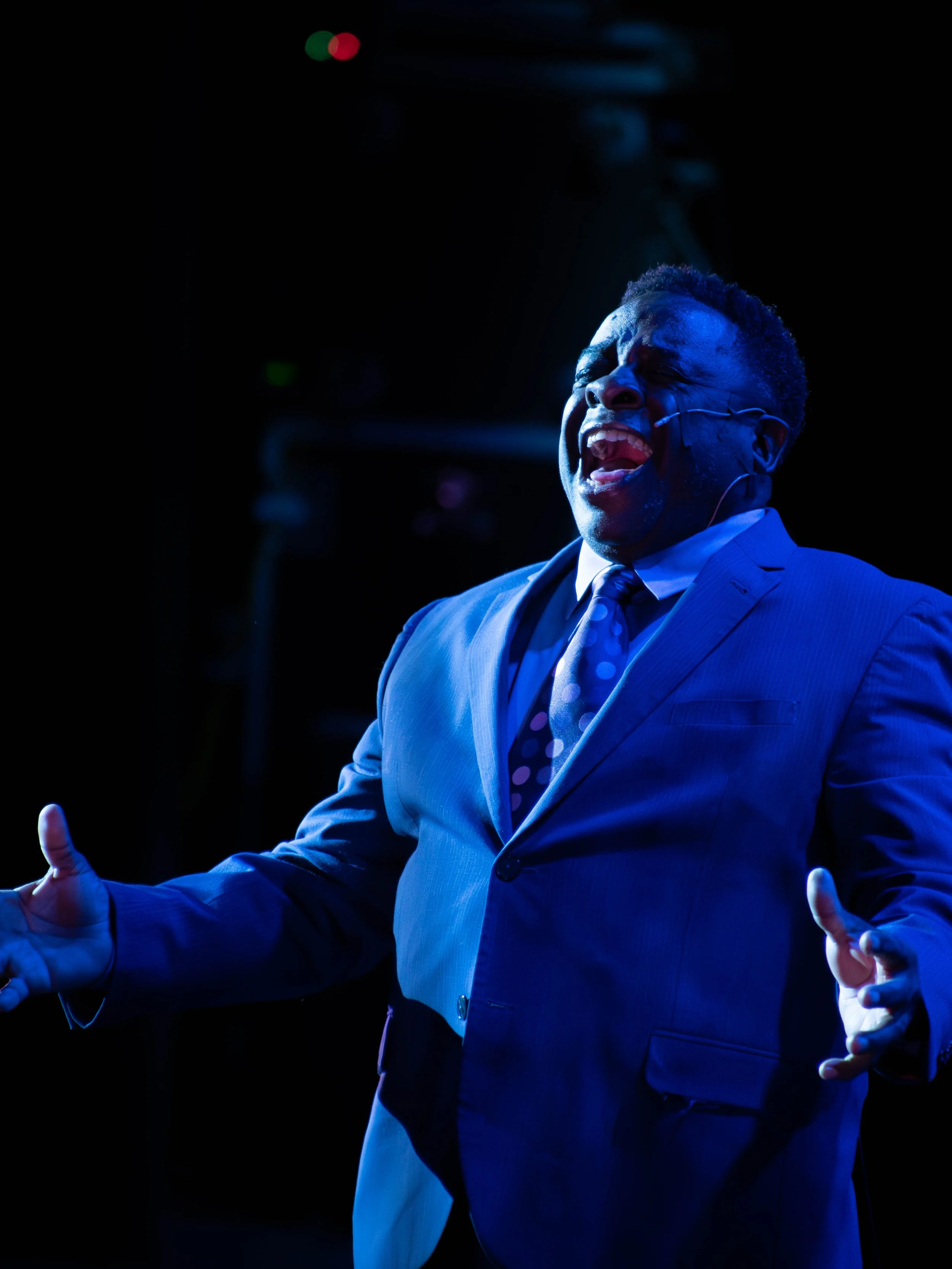 A man in a blue suit and polka dot tie passionately speaking or singing on stage, with expressive gestures and emotional facial expression, illuminated by stage lighting.