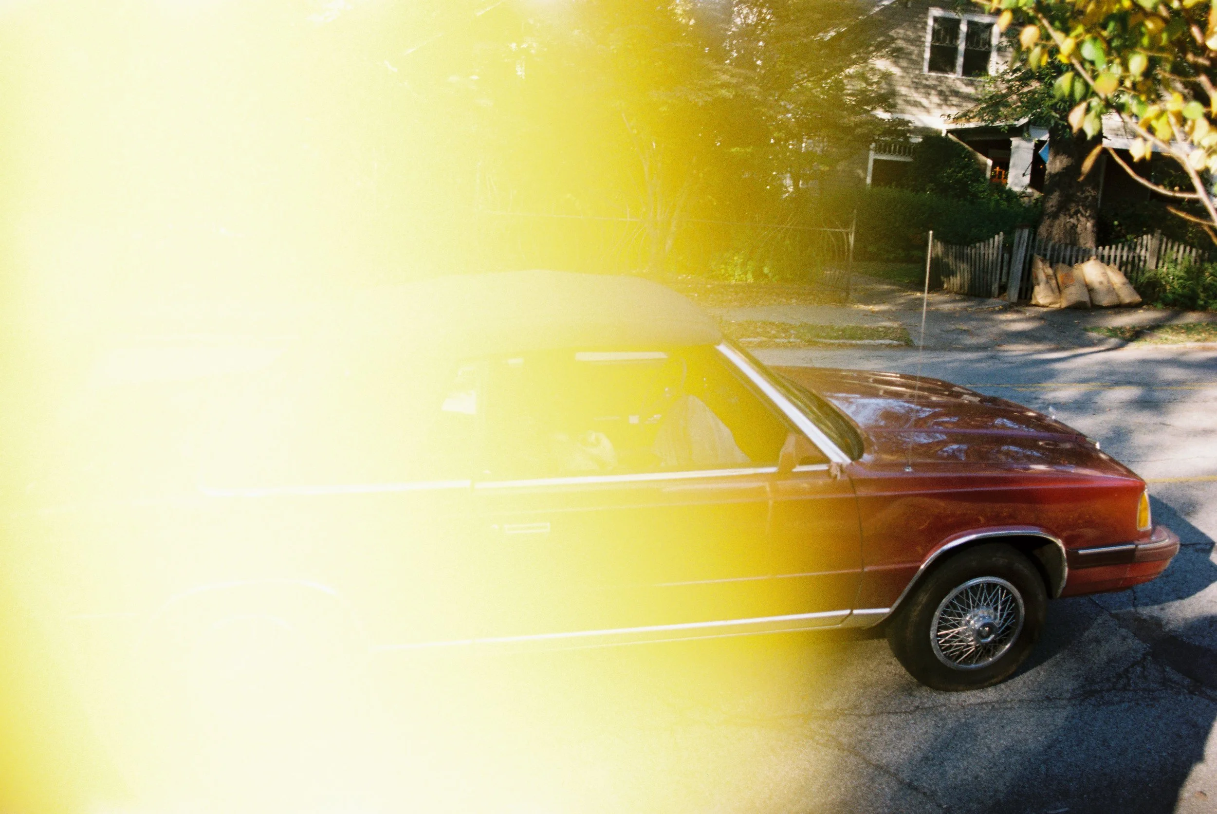 A vintage red car parked on a residential street with trees and houses in the background, partially obscured by a large yellowish light streak.