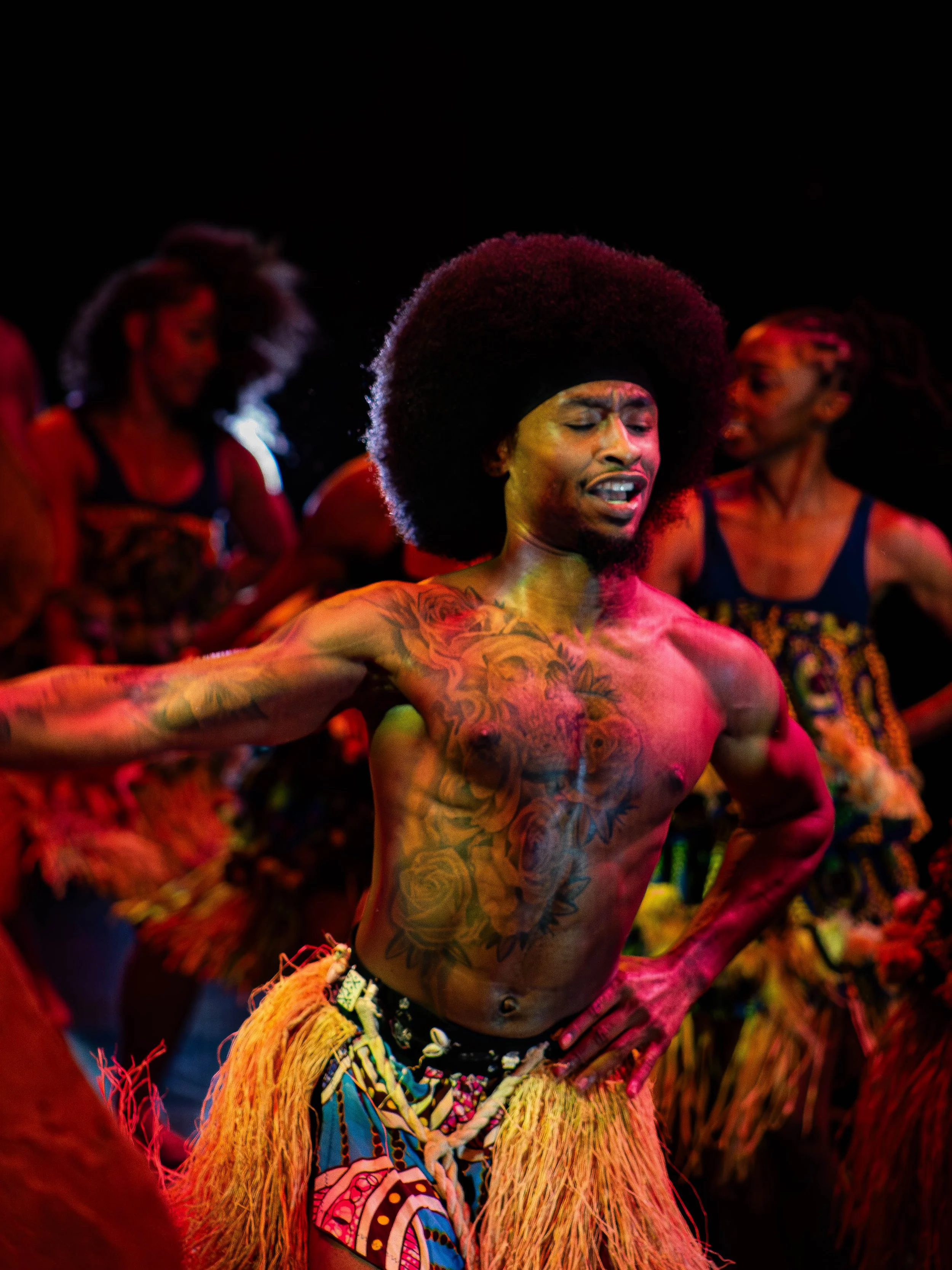 A shirtless man with a large afro hairstyle and extensive tattoos practices traditional dance with a group of women in colorful costumes.