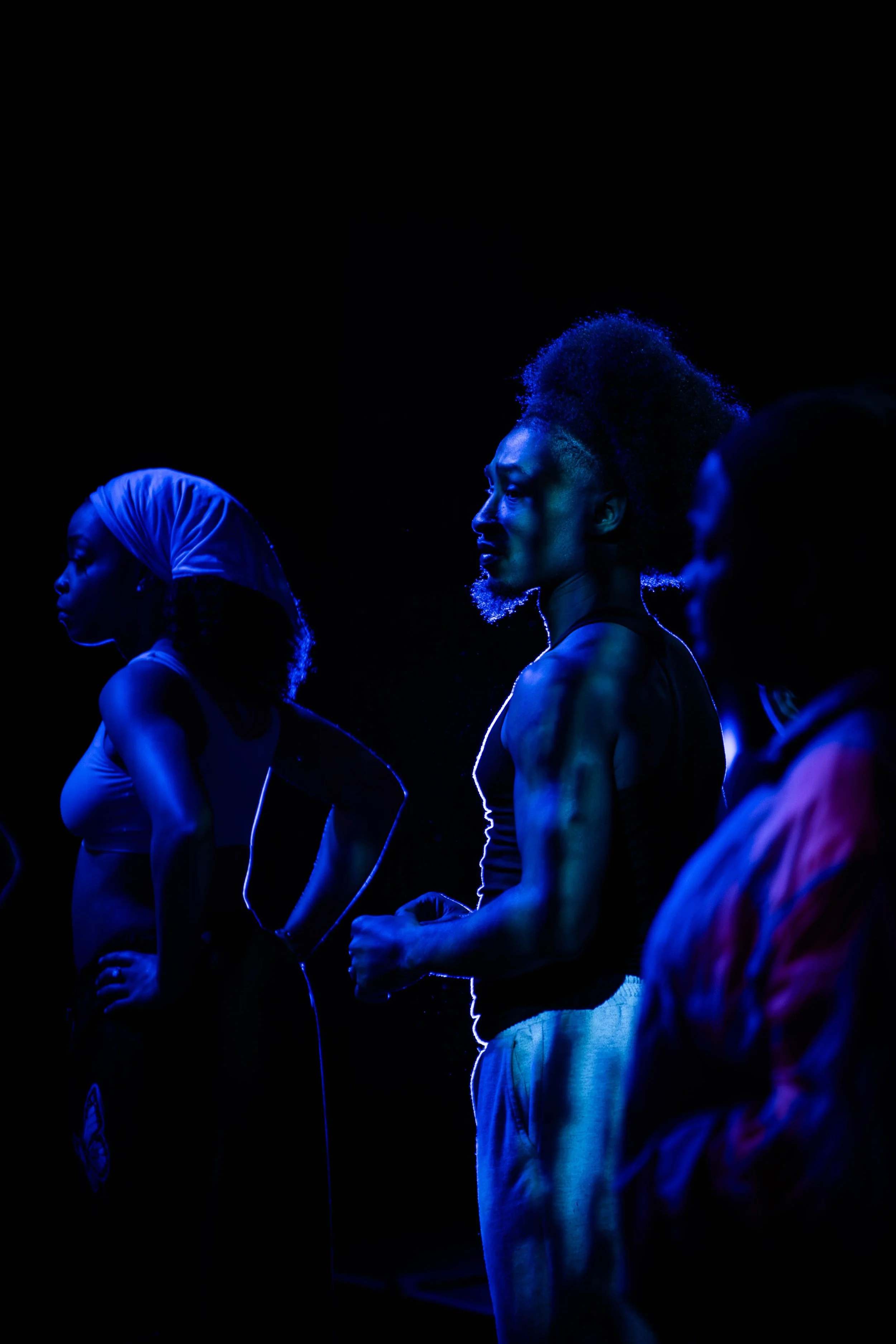 Group of diverse women with muscular physiques standing in a line, illuminated with blue lighting, showing intense expressions and athletic clothing, possibly in a fitness or dance setting.