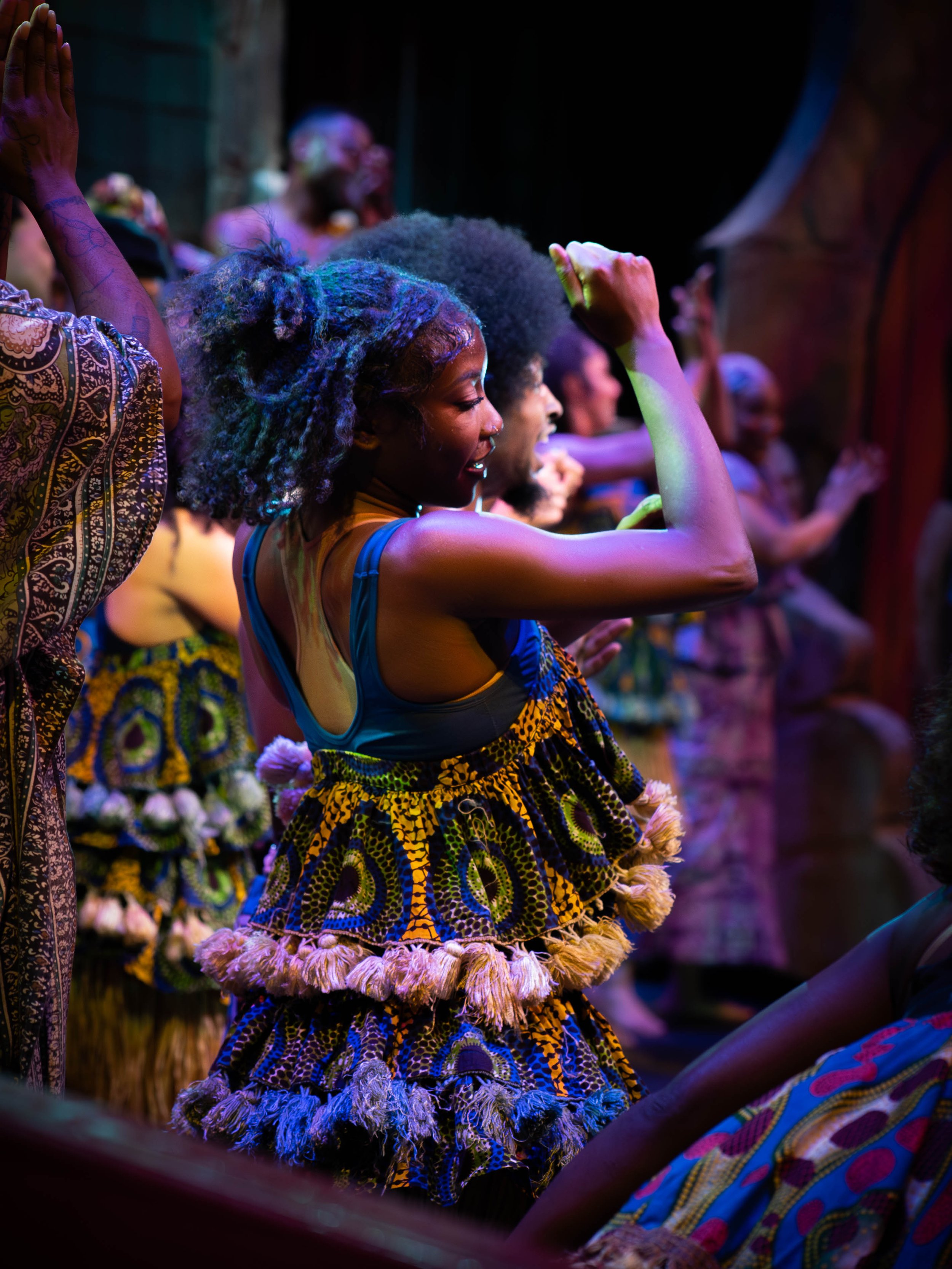 Group of people dancing and celebrating in colorful traditional African clothing under vibrant lighting.