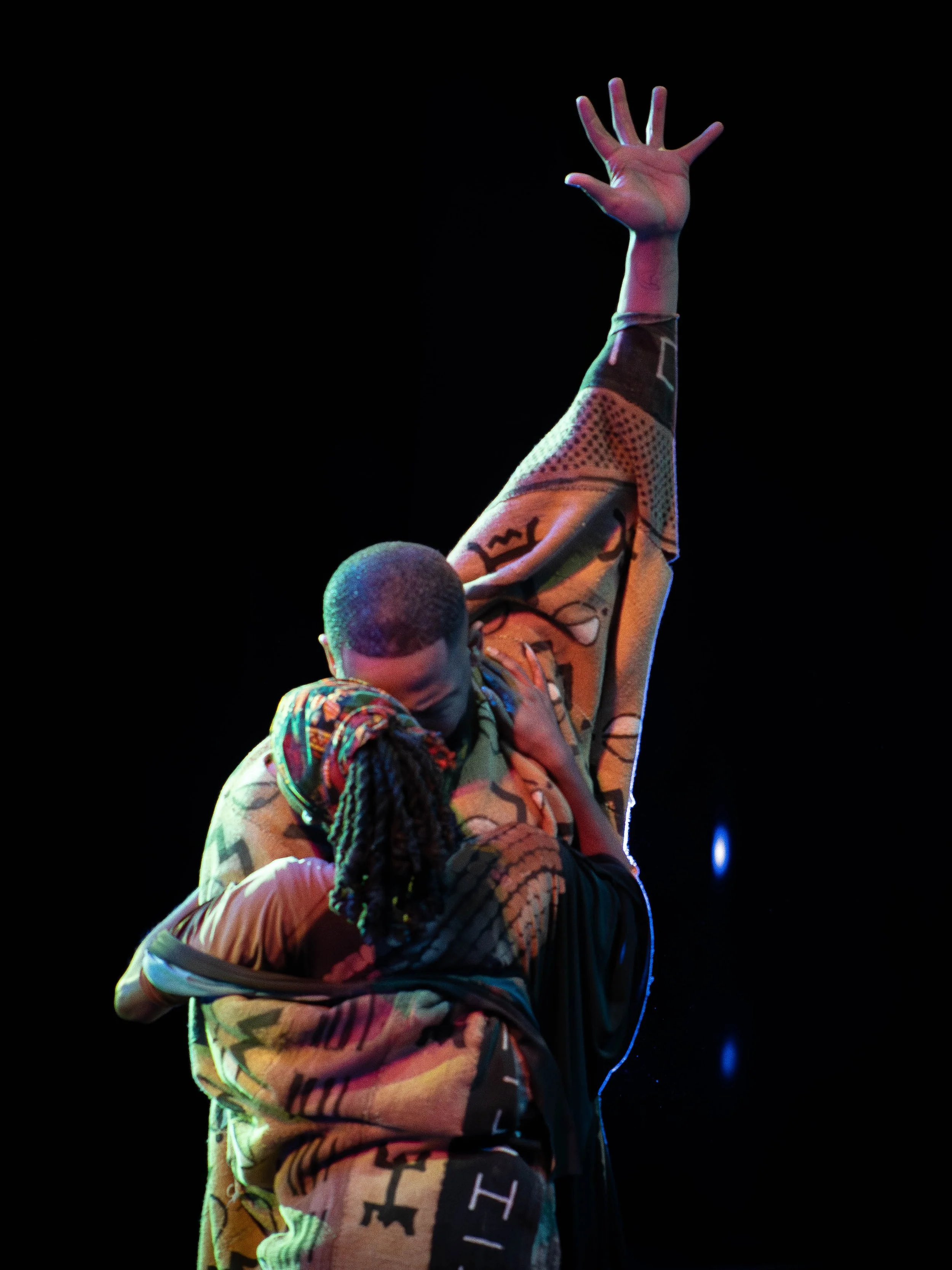 Two dancers performing on stage, one with an arm raised and the other with head lowered, both wearing colorful patterned clothing, against a dark background.