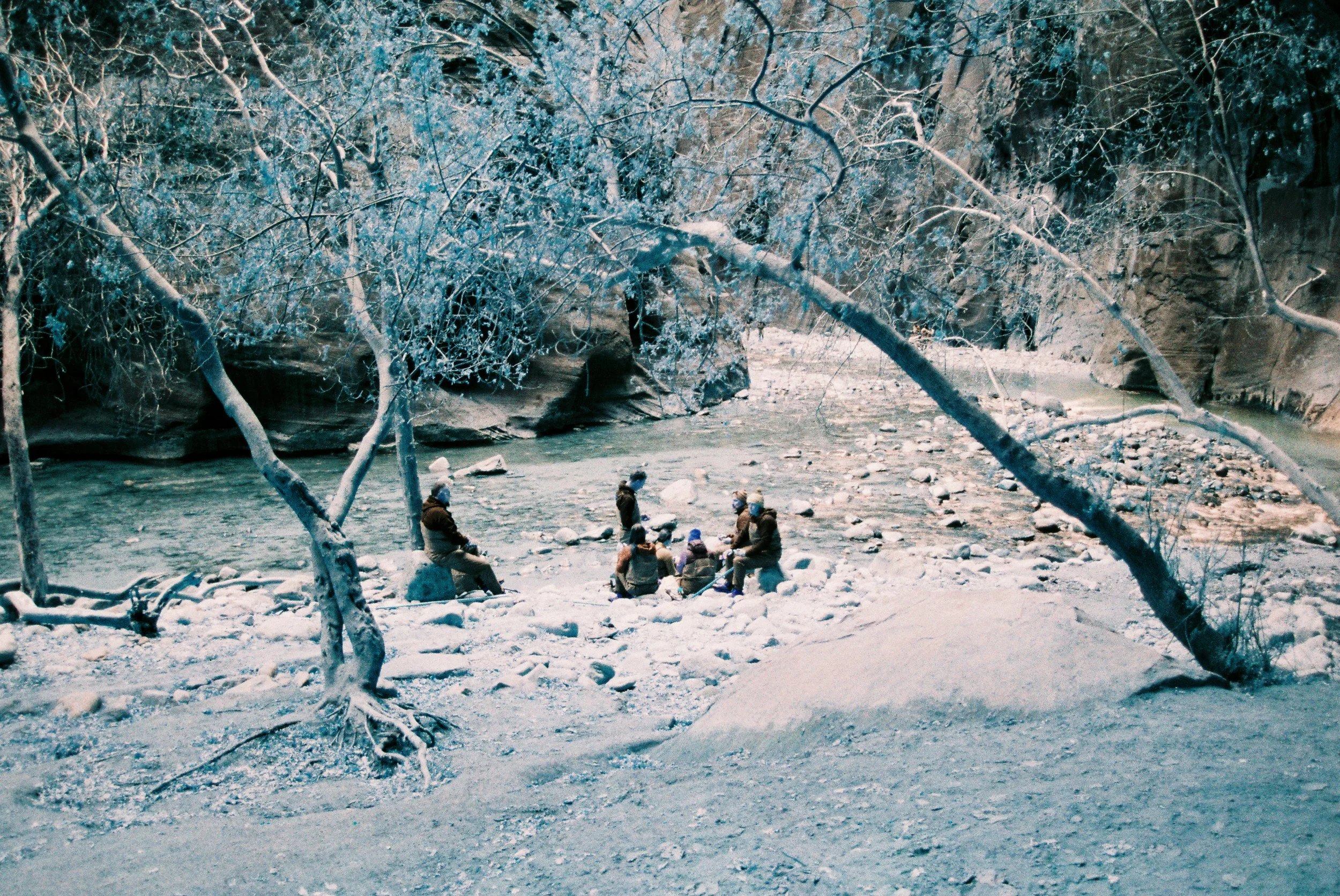 A group of people sitting and standing by a riverbank in a snowy forest, with snow-covered trees and rocks surrounding them.